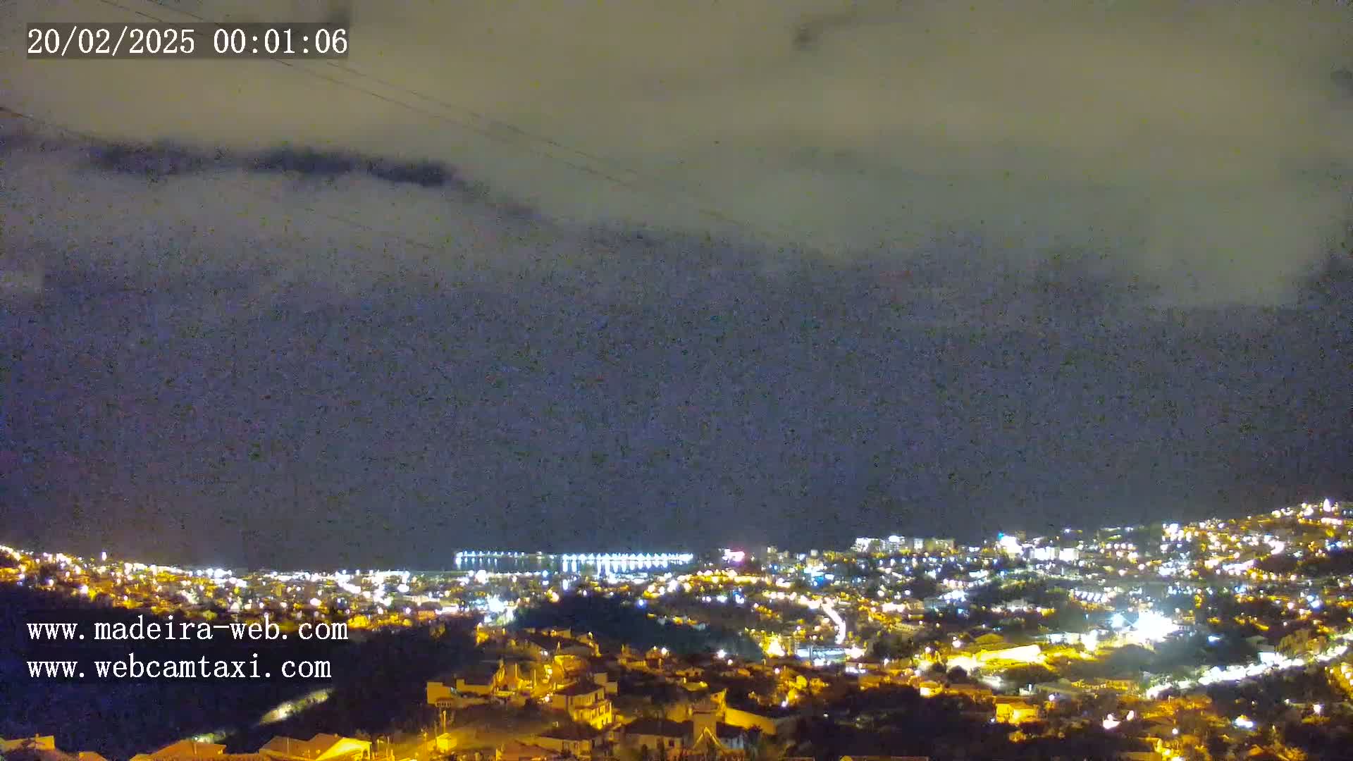 A nighttime, cloudy view of a city illuminated by lights, stretching down a hillside toward a body of water.