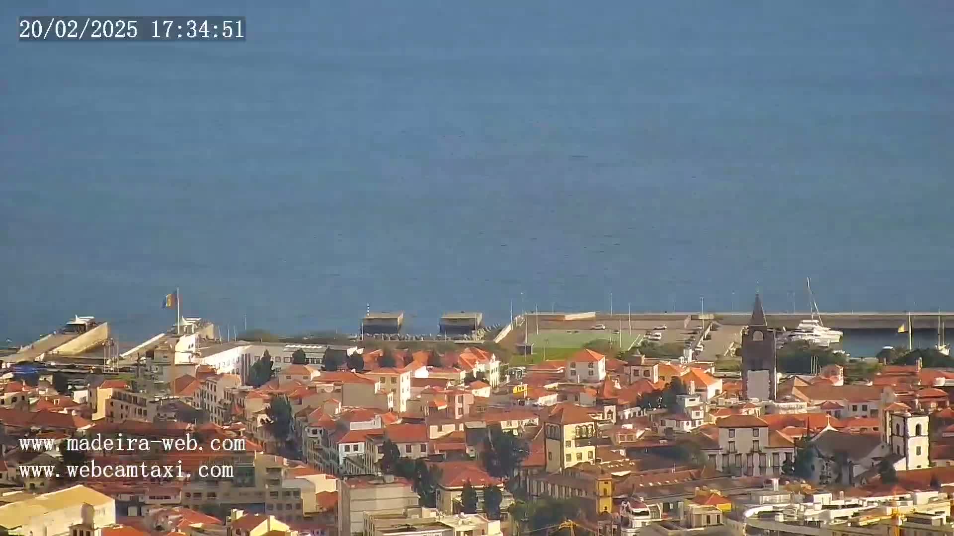 A coastal town with red-tiled roofs and a tall church tower is situated next to a calm, blue ocean under a clear sky.