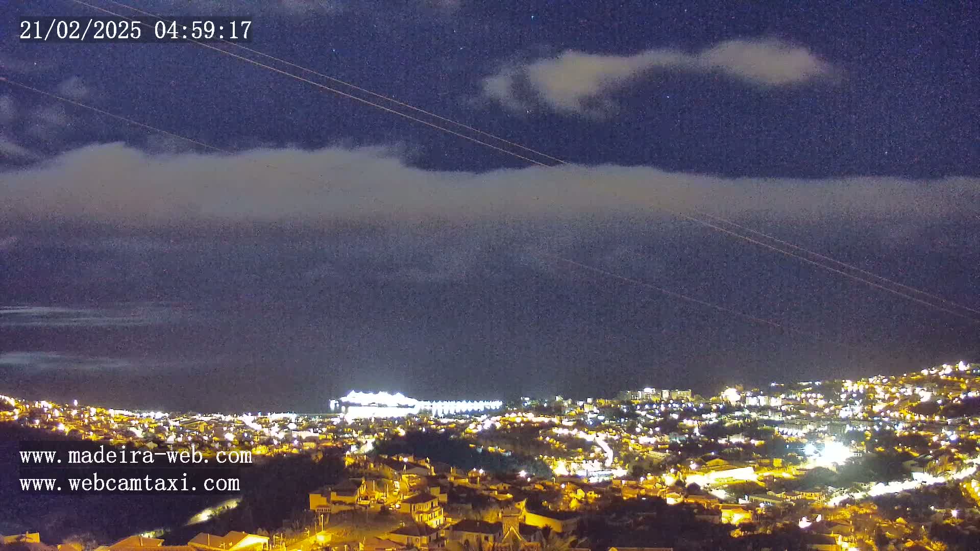 A nighttime, low-cloud-covered view of a city illuminated by lights, extending to a coastline under a dark, starry sky.