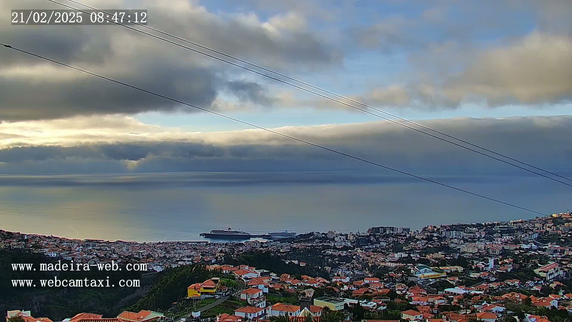 A coastal town with red-roofed houses stretches along the ocean under a partly cloudy sky.