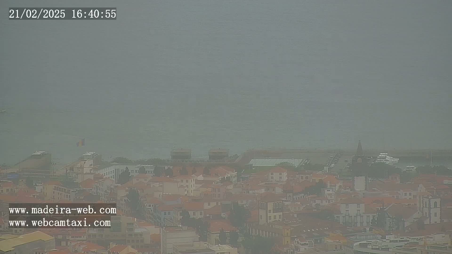 A hazy view of a coastal town with red-roofed buildings, a body of water, and a distant pier is visible under a gray sky.