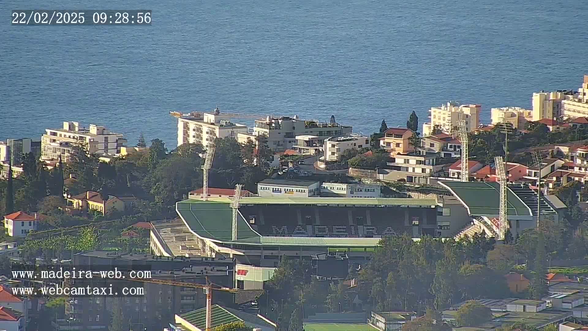 A coastal town with buildings and a stadium is visible on a clear day, with calm blue water in the background.