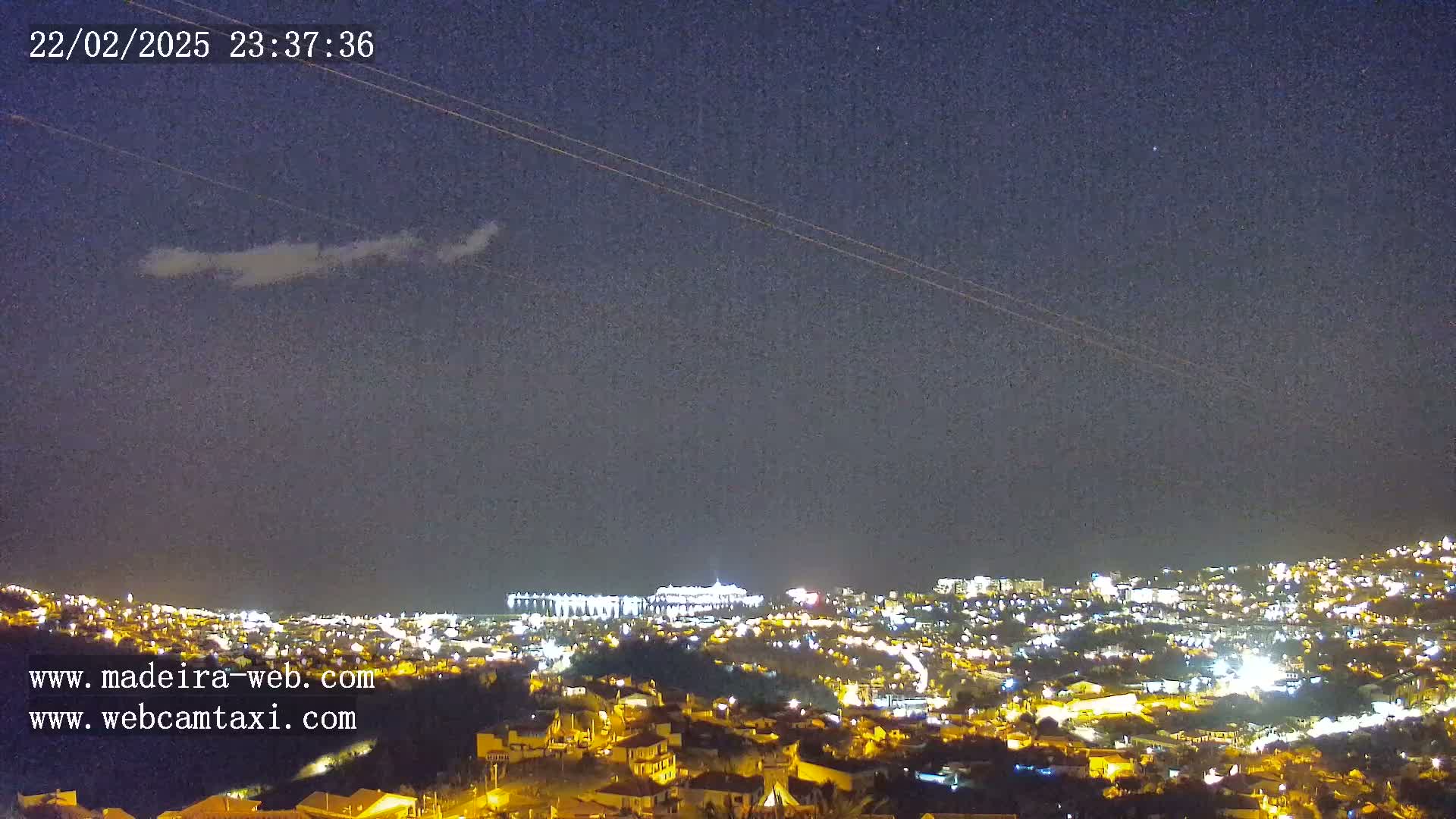 A nighttime view of a city illuminated by lights under a mostly clear, dark sky with a few wispy clouds.