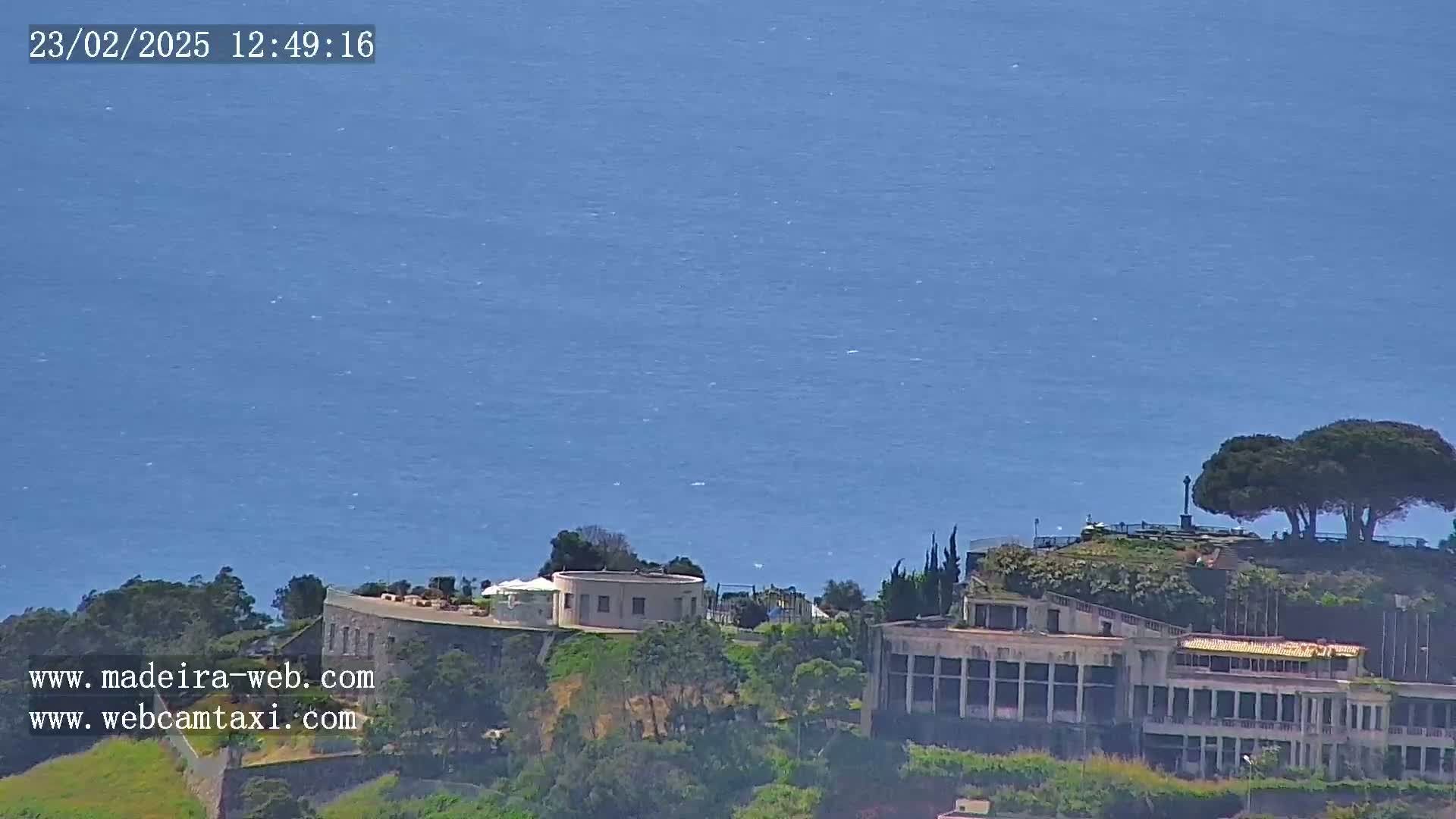 A coastal scene on a clear day shows a large building complex overlooking a calm blue ocean, with some trees and green vegetation in the foreground.