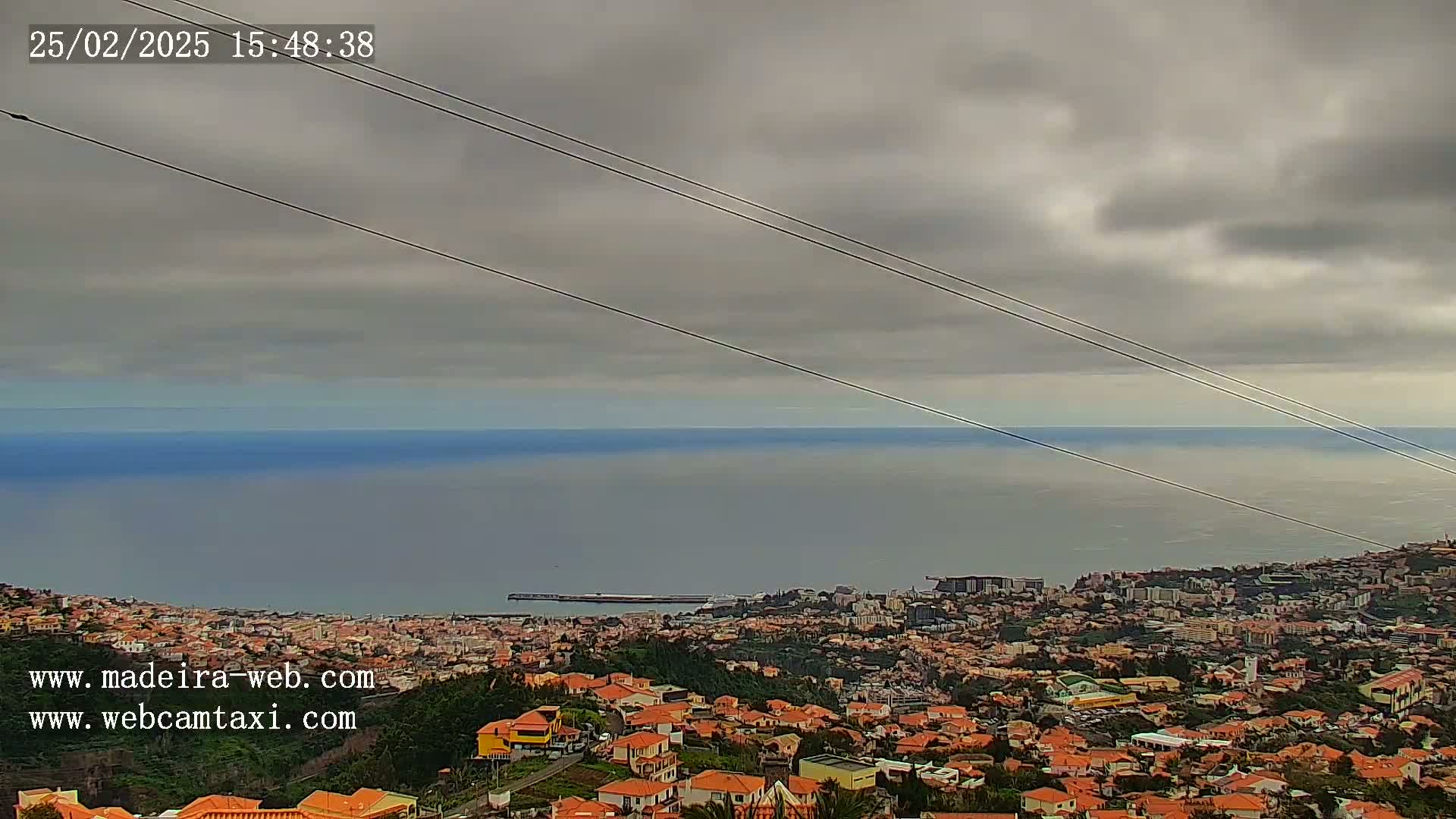 A coastal city with orange-roofed buildings stretches to a calm ocean under a mostly cloudy sky.