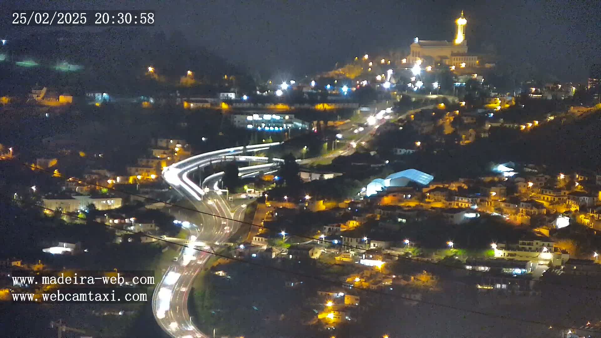 A nighttime, high-angle view of a town situated on a hillside, with illuminated buildings, roads, and traffic, featuring a prominent structure atop a hill.