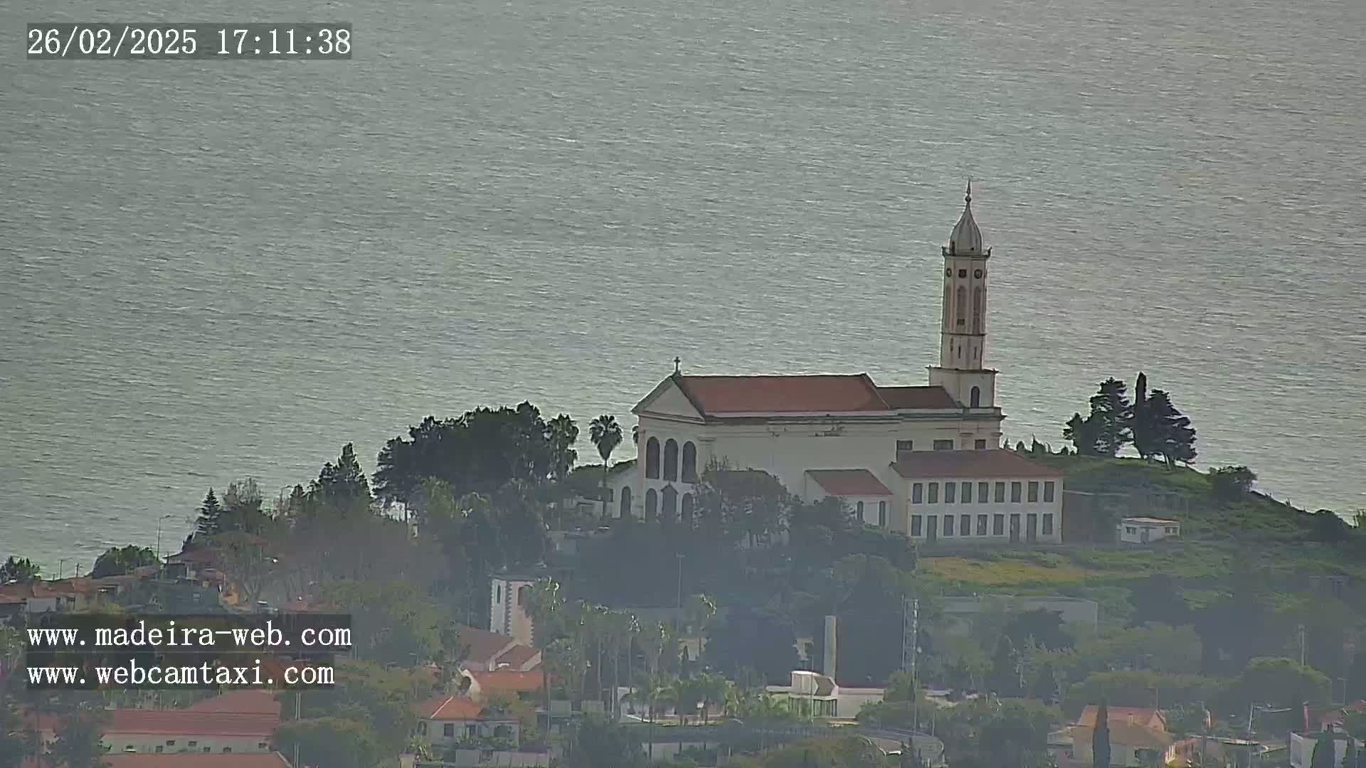 A white church with a tall tower sits atop a small hill overlooking a calm body of water, surrounded by green vegetation and other buildings.