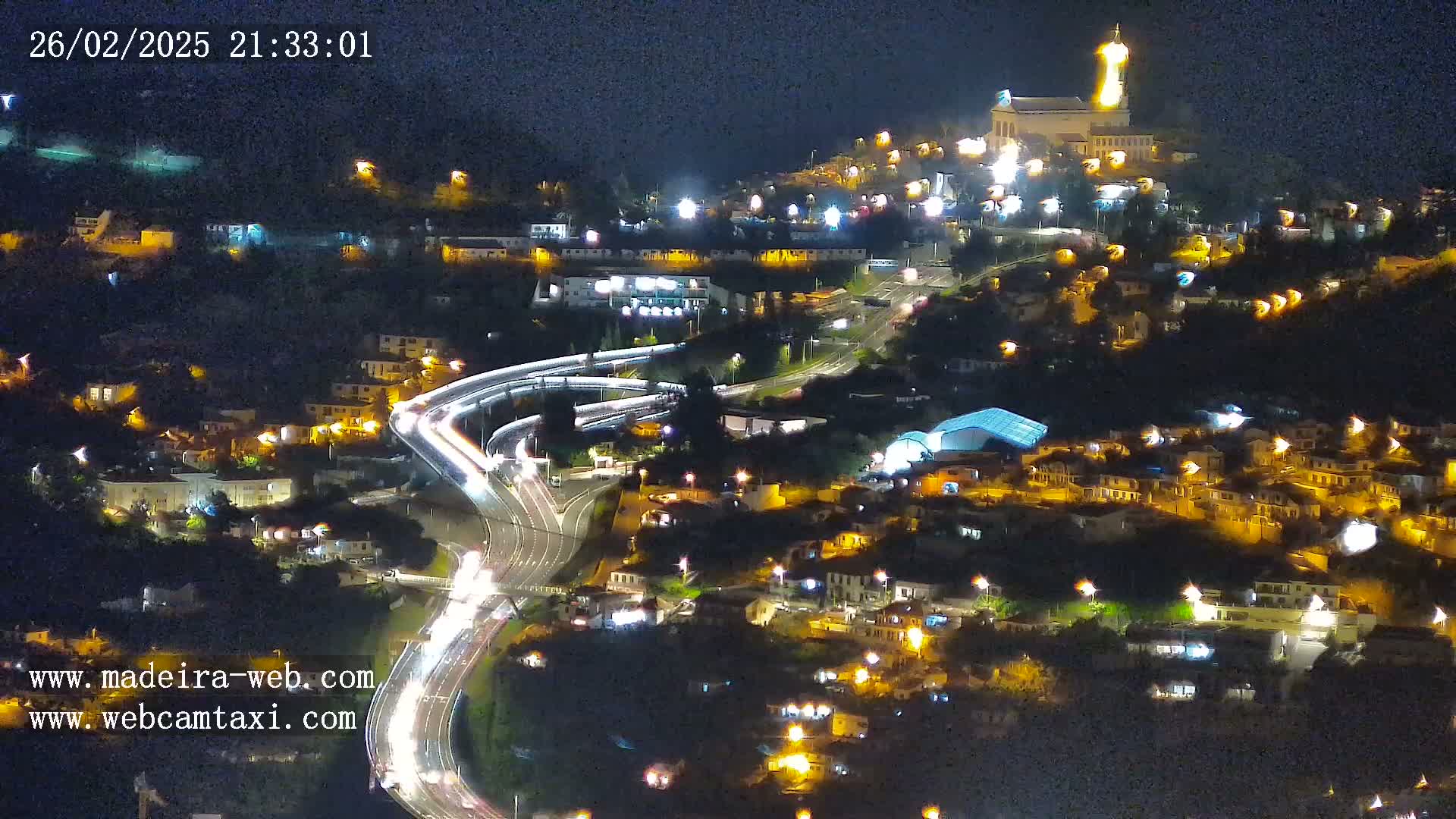 A nighttime aerial view of a town on a hillside, with a brightly lit church atop the hill and traffic flowing on roads below.