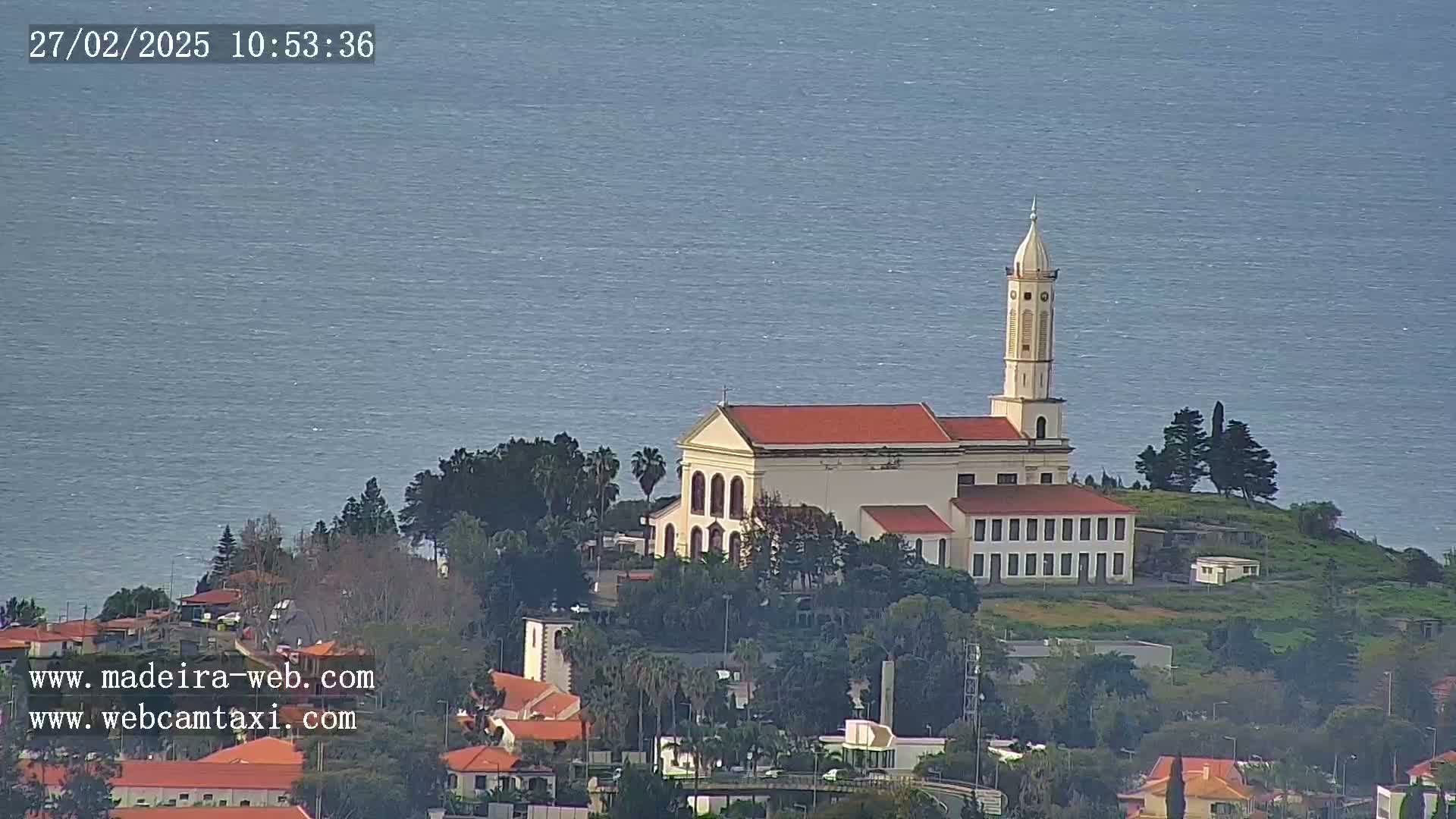 A church with a red roof and tall bell tower sits atop a hill overlooking a calm ocean on a clear day.