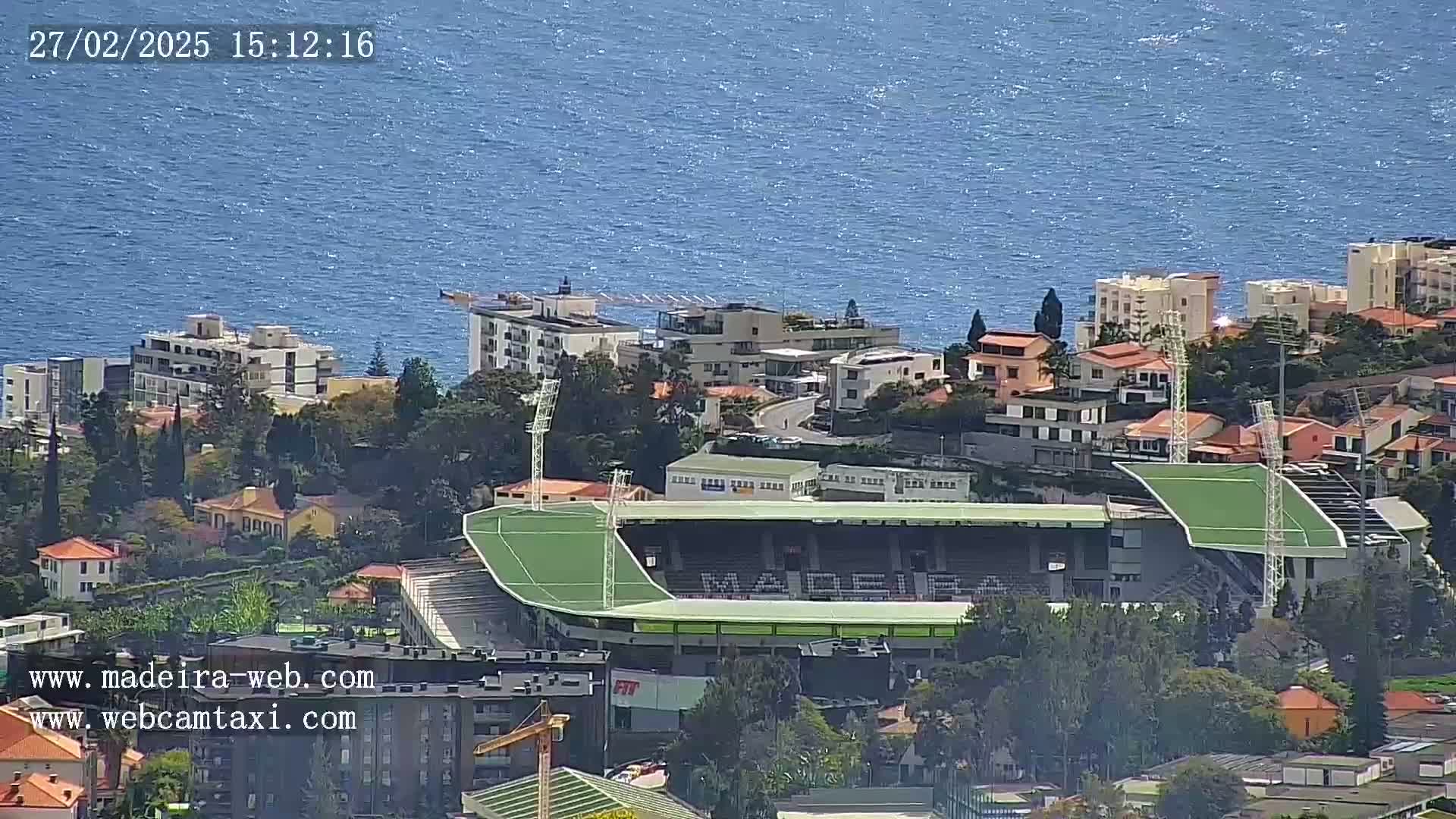 A soccer stadium with a green field is nestled amongst buildings near a body of blue water on a sunny day.