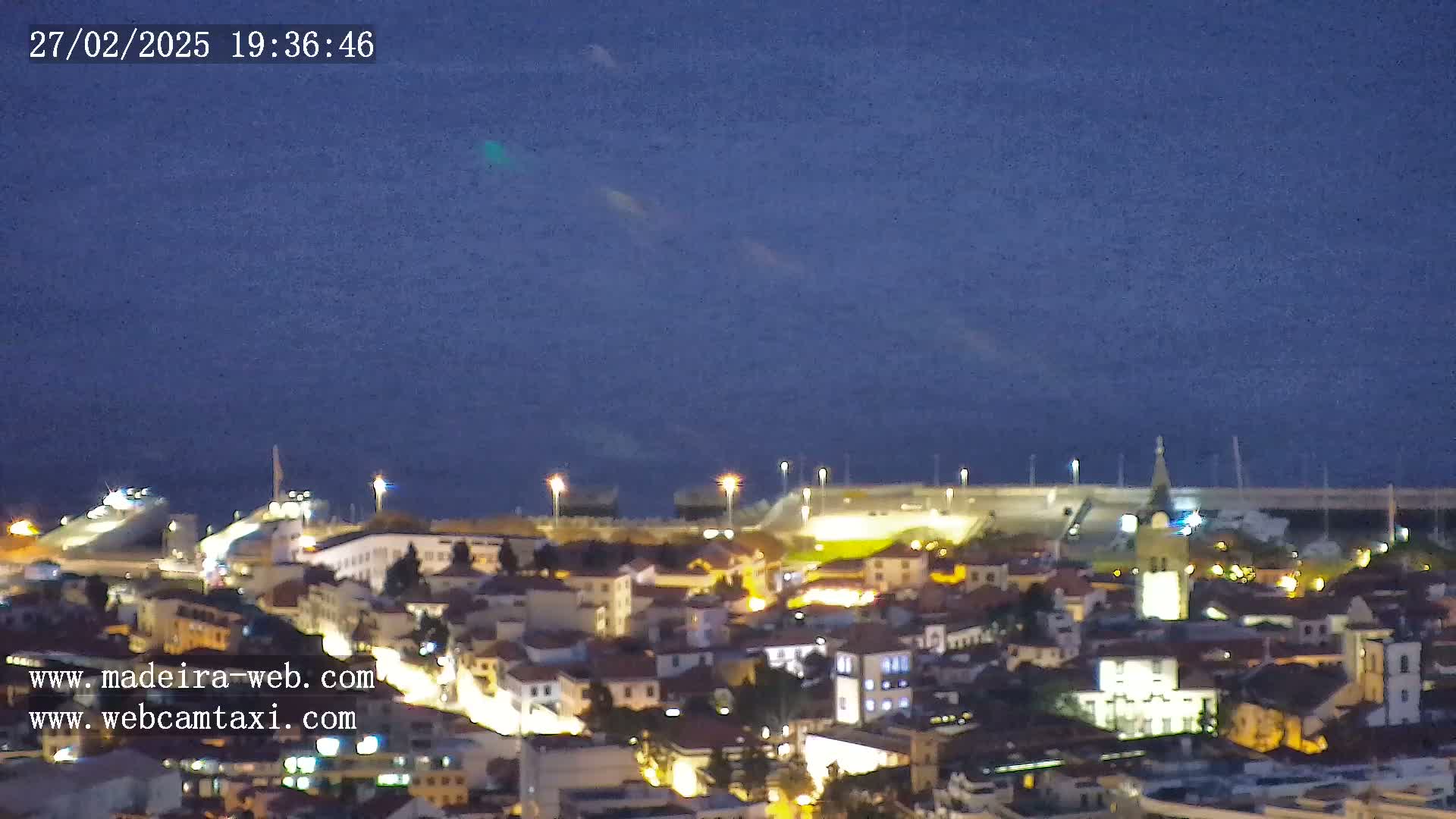 A nighttime, aerial view of a coastal town with a harbor, illuminated buildings, and a dark blue sky.