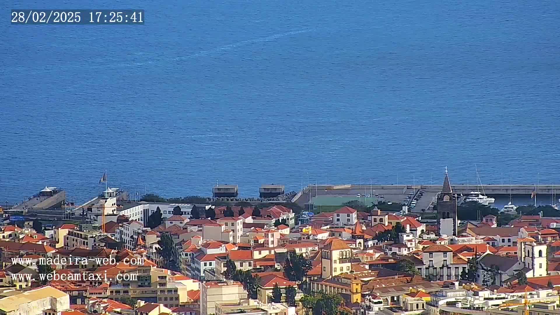 A coastal town with red-roofed buildings stretches toward a calm blue ocean on a sunny day.