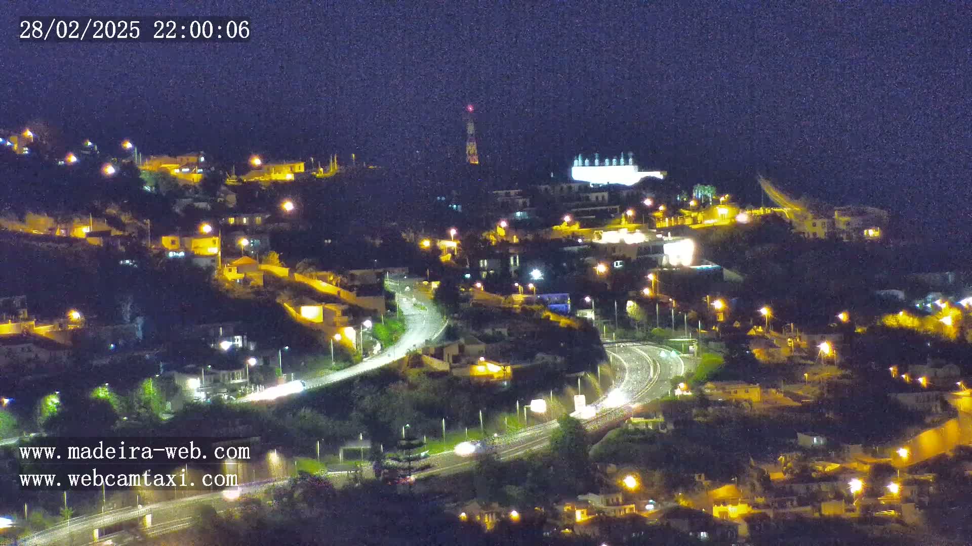 A nighttime, high-angle view shows a hilly landscape with illuminated houses and roads, with some vehicle lights visible on the roads.
