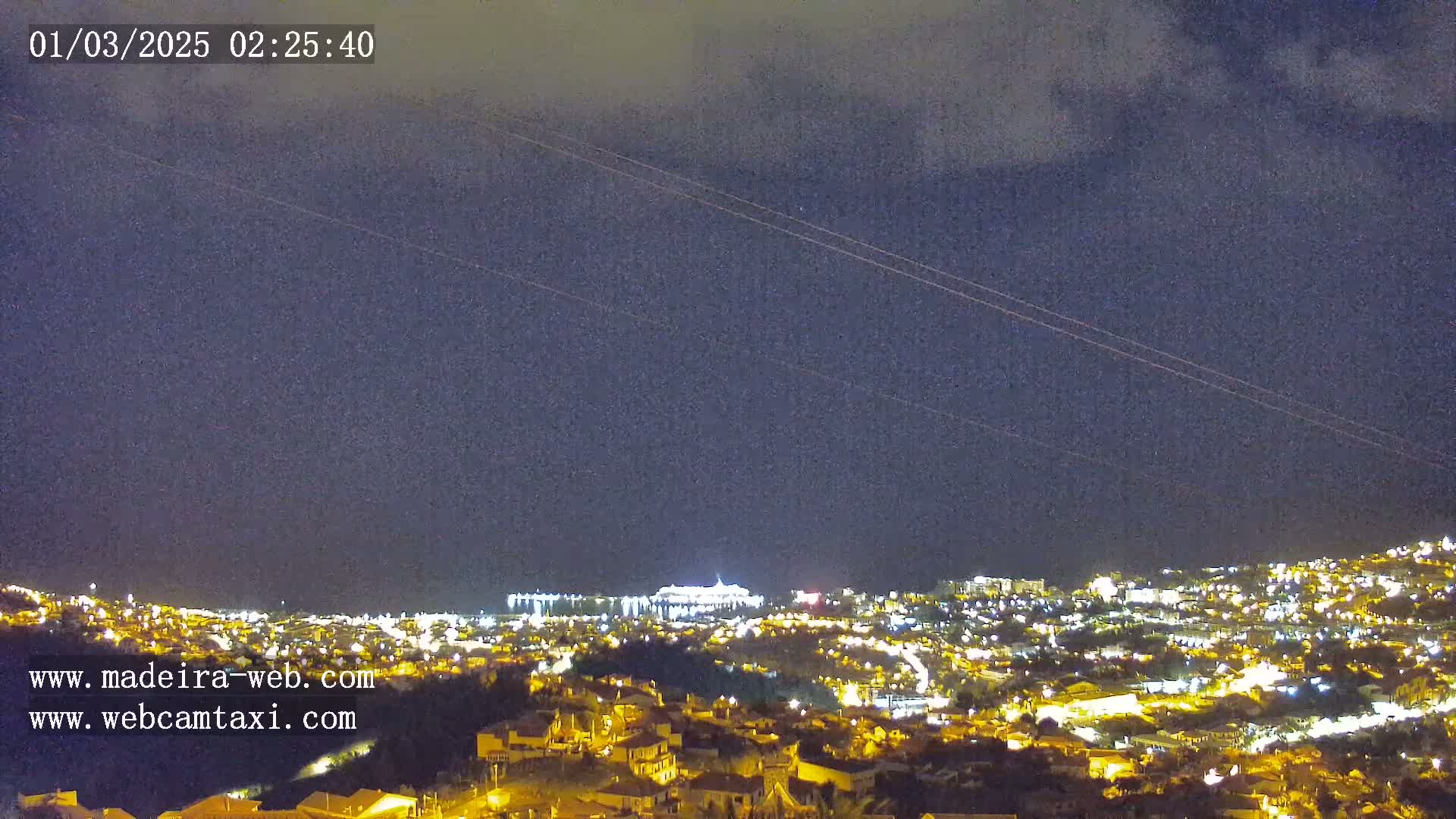 A nighttime, mostly cloudy view overlooking a city illuminated by streetlights, with a larger structure brightly lit in the distance.