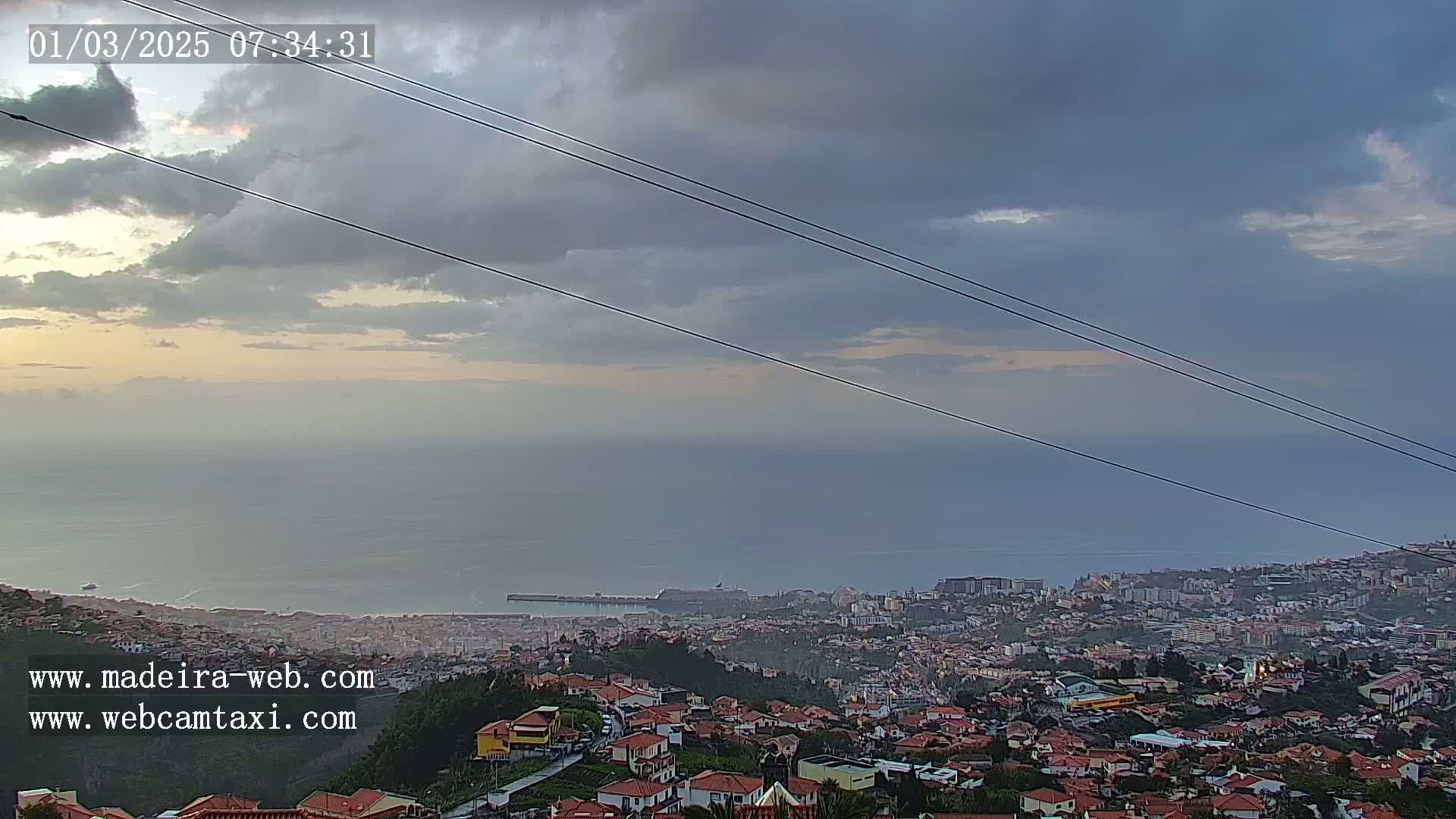 A coastal city with red-roofed houses sprawls below a cloudy sky, overlooking a calm ocean.