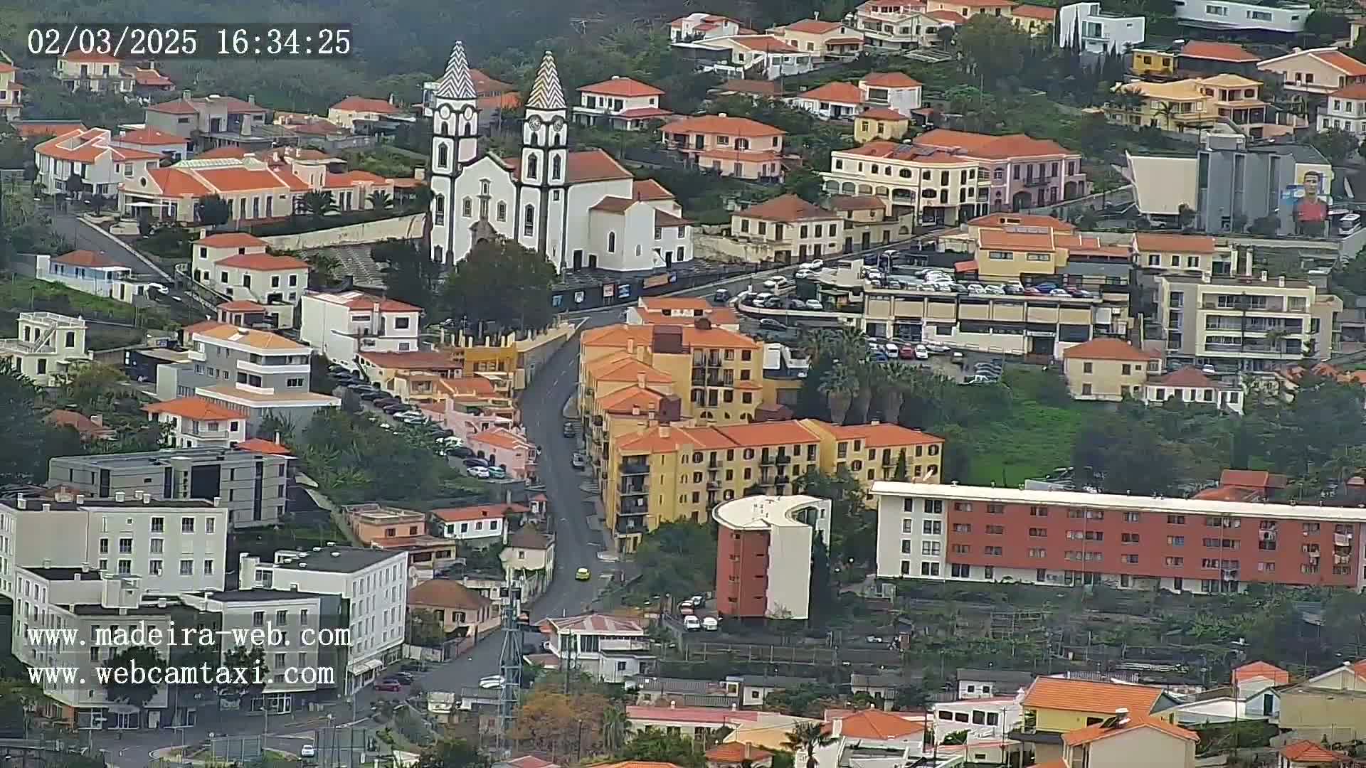 An aerial view shows a town with predominantly red-roofed buildings, a prominent white church with twin towers, and overcast skies.