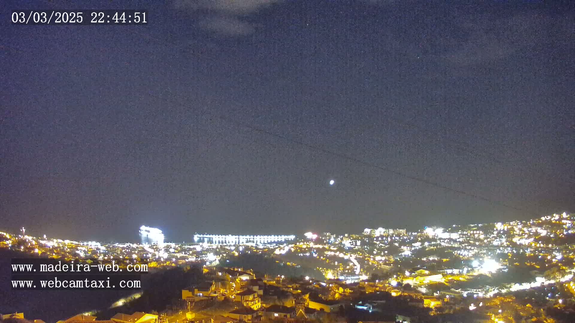 A nighttime view of a city illuminated by lights, with a clear sky showing a few stars and a bright celestial body.