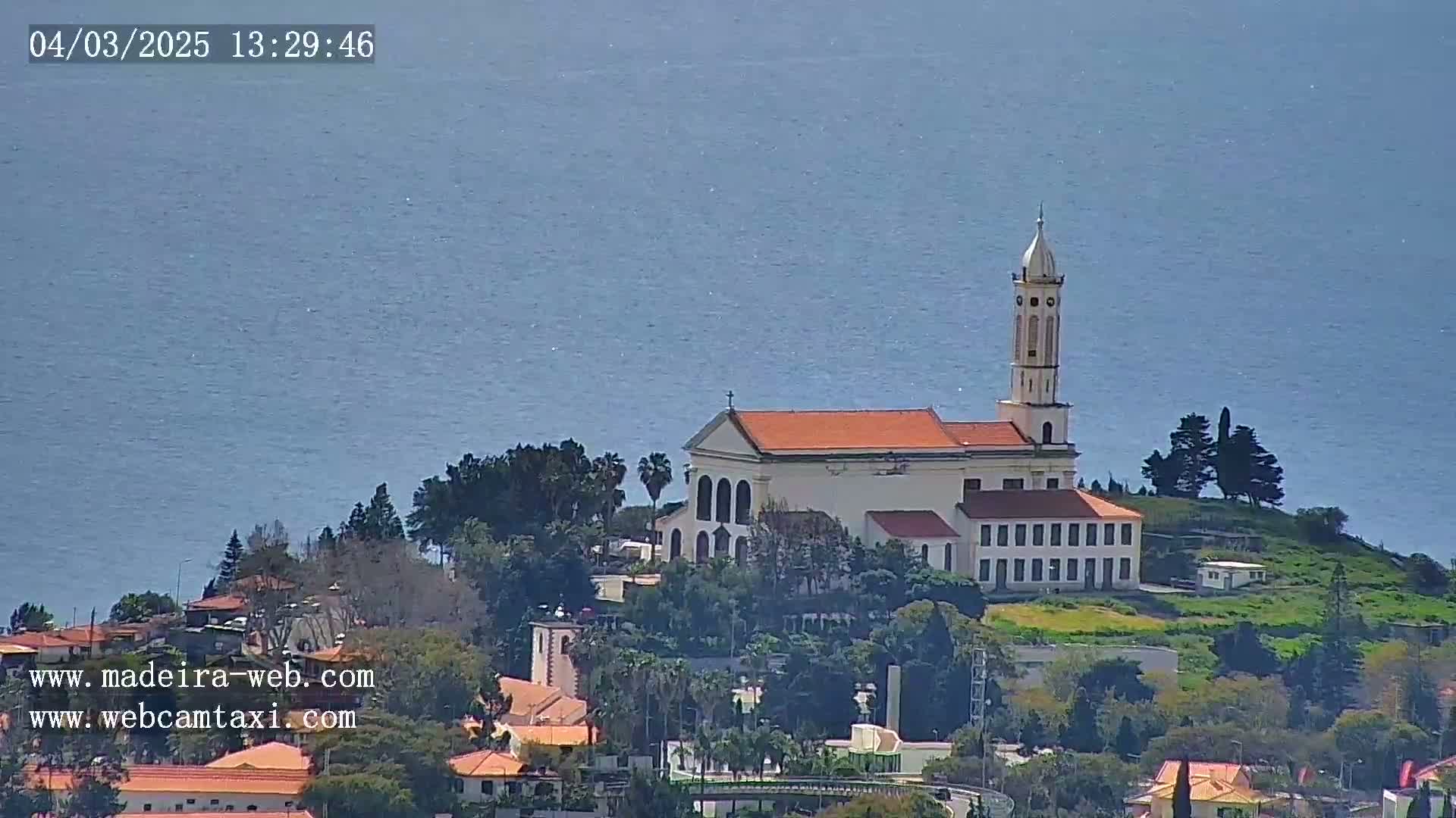 A white church with an orange roof and tall steeple sits atop a hill overlooking a calm ocean on a clear day.
