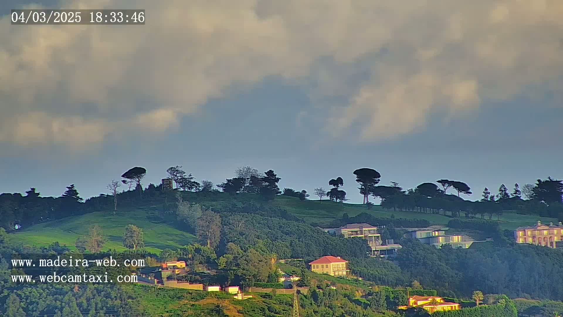 A partly cloudy sky overlooks a verdant hillside dotted with trees and houses.