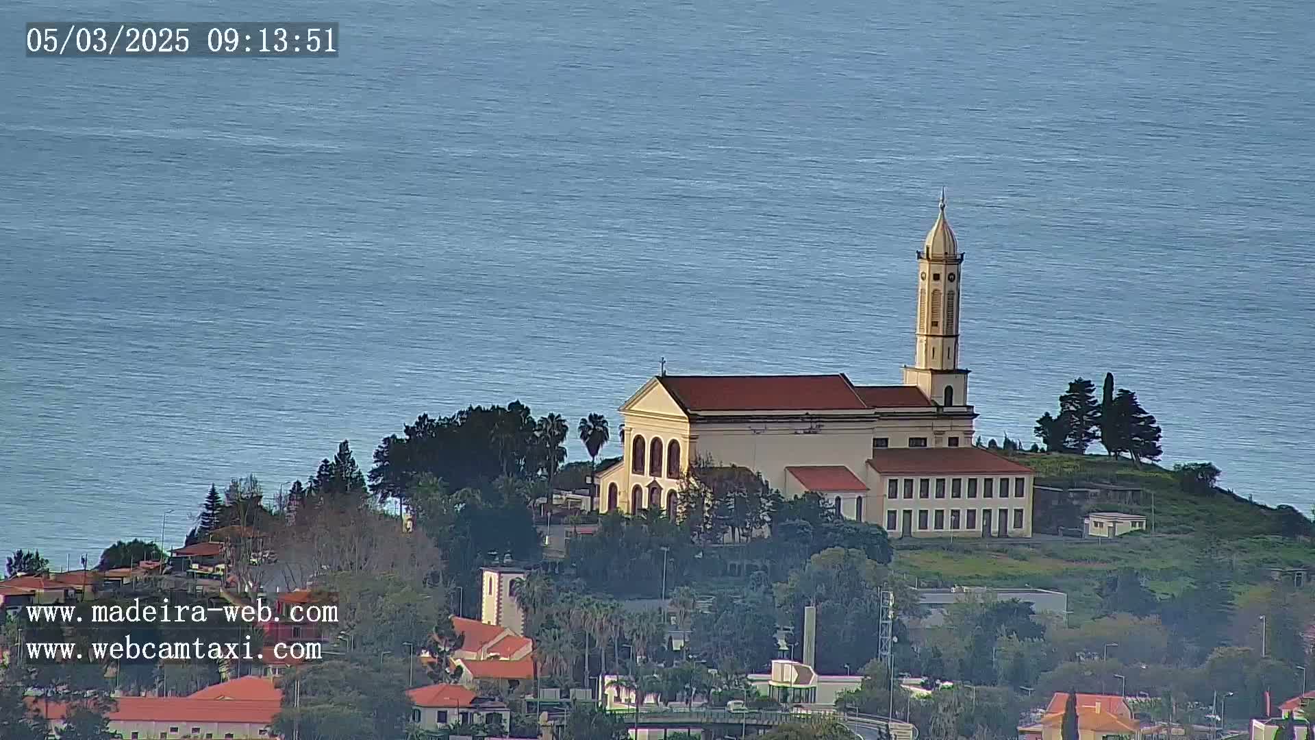 A church with a tall bell tower sits atop a hill overlooking a calm ocean.