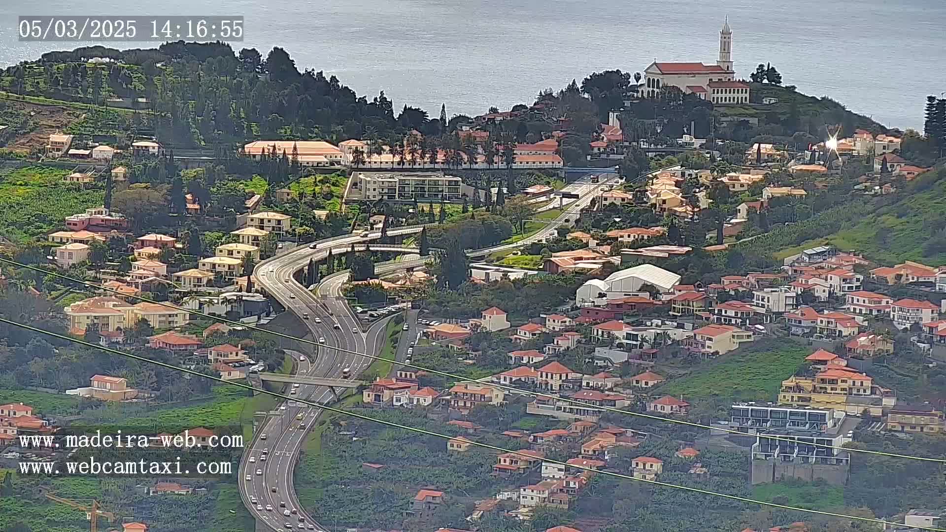 A high-angle view shows a coastal town with a church on a hill, a highway weaving through residential areas, and a clear, sunny day.