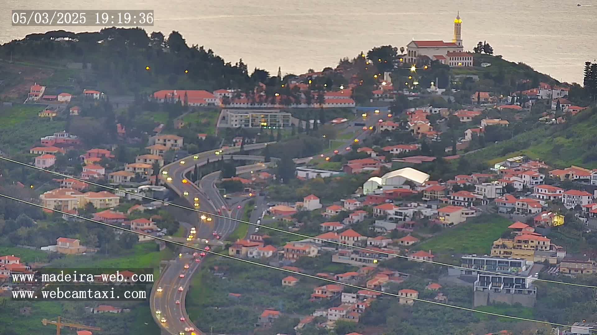 An aerial view shows a coastal town at dusk with a church on a hill overlooking a highway with cars driving on it.
