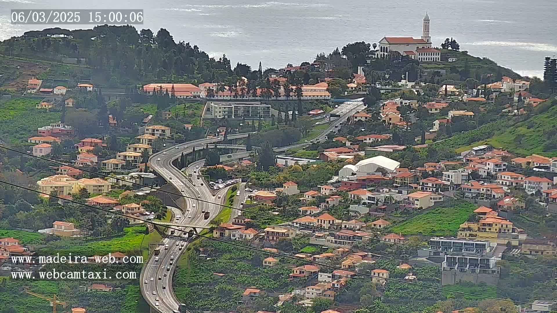 An aerial view of a coastal town with a highway winding through it, under a mostly cloudy sky with a body of water visible in the background.