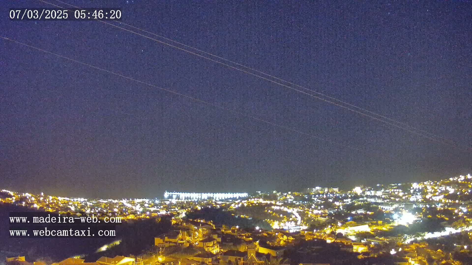 A nighttime view of a city illuminated by lights under a clear, starry sky with two faint contrails.