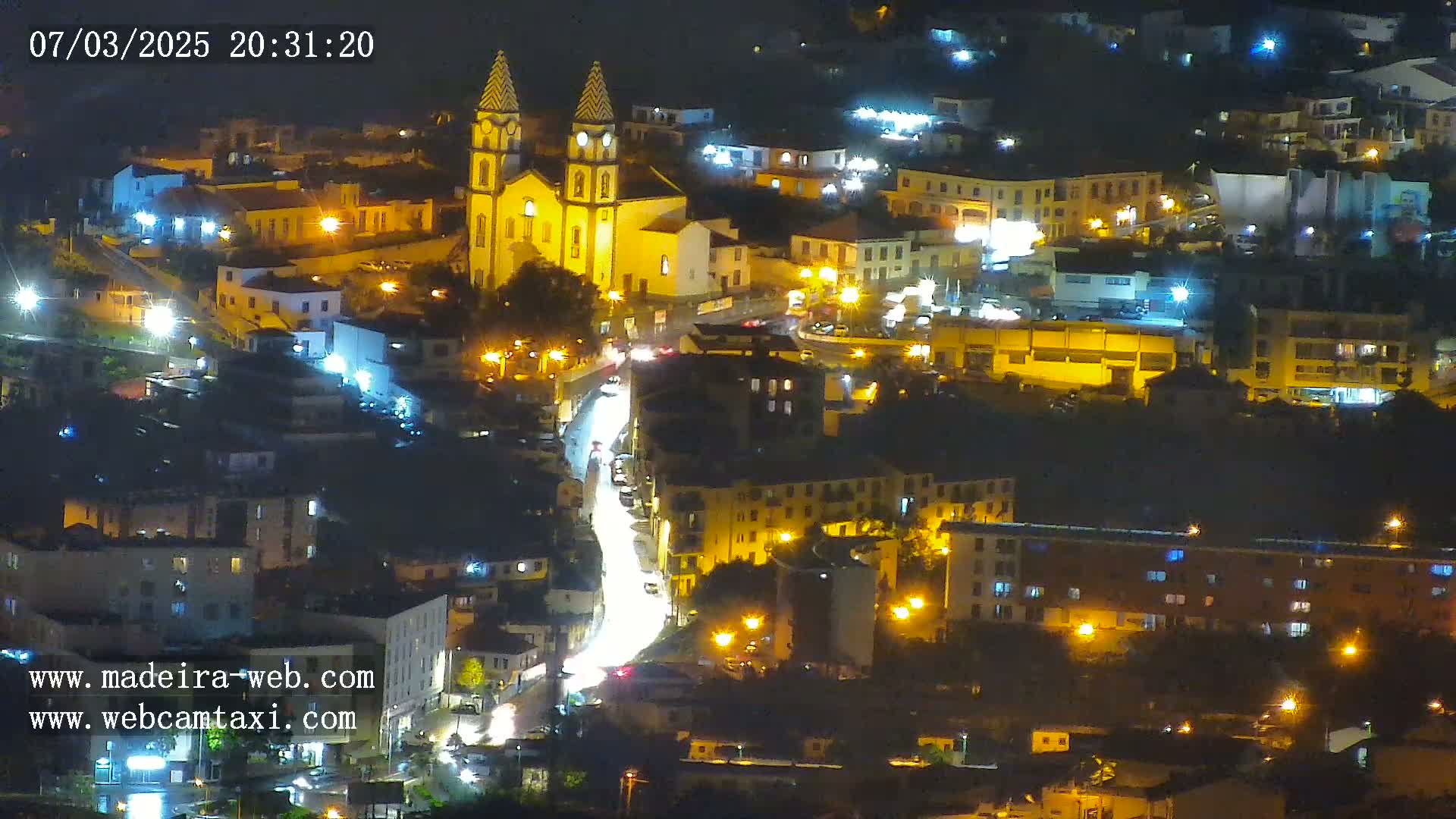 A nighttime, aerial view of a town illuminated by streetlights, featuring a prominent church at its center and a brightly lit street winding through the buildings.