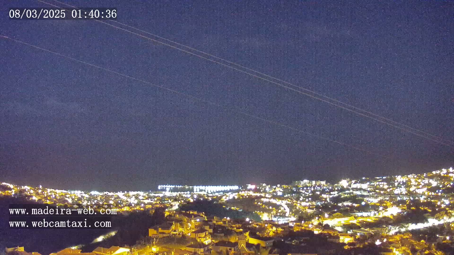 A nighttime view of a city illuminated by lights under a dark, clear sky with faint streaks possibly from airplanes.