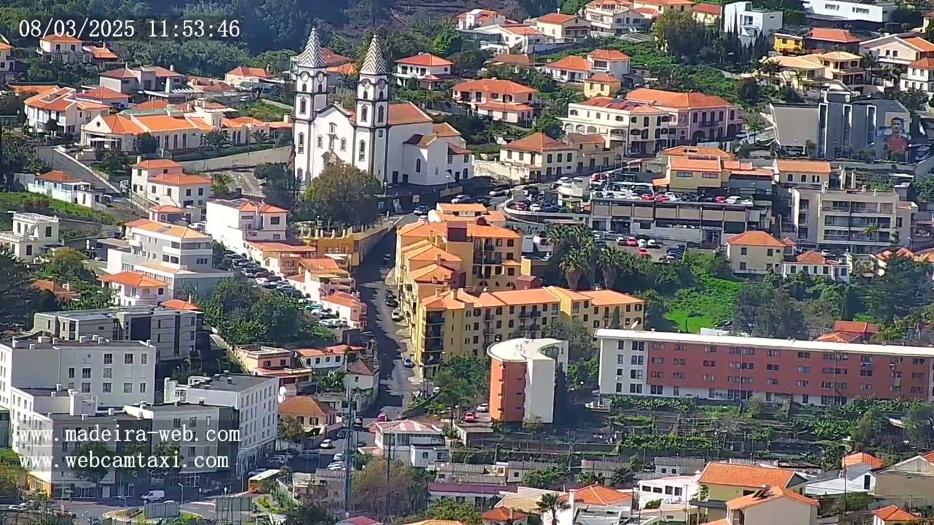 A sunlit aerial view of a town with numerous orange-roofed buildings, a prominent white church, and a curved apartment building.