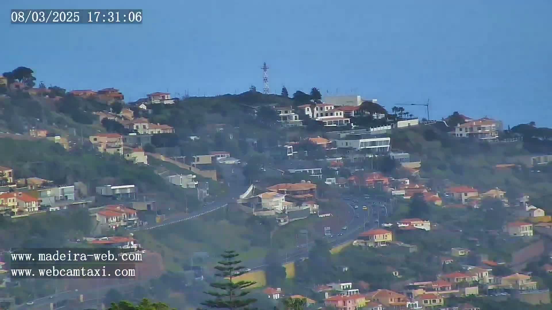 A hazy view shows a hillside densely covered with houses, a road winding through, and a communications tower atop the hill.
