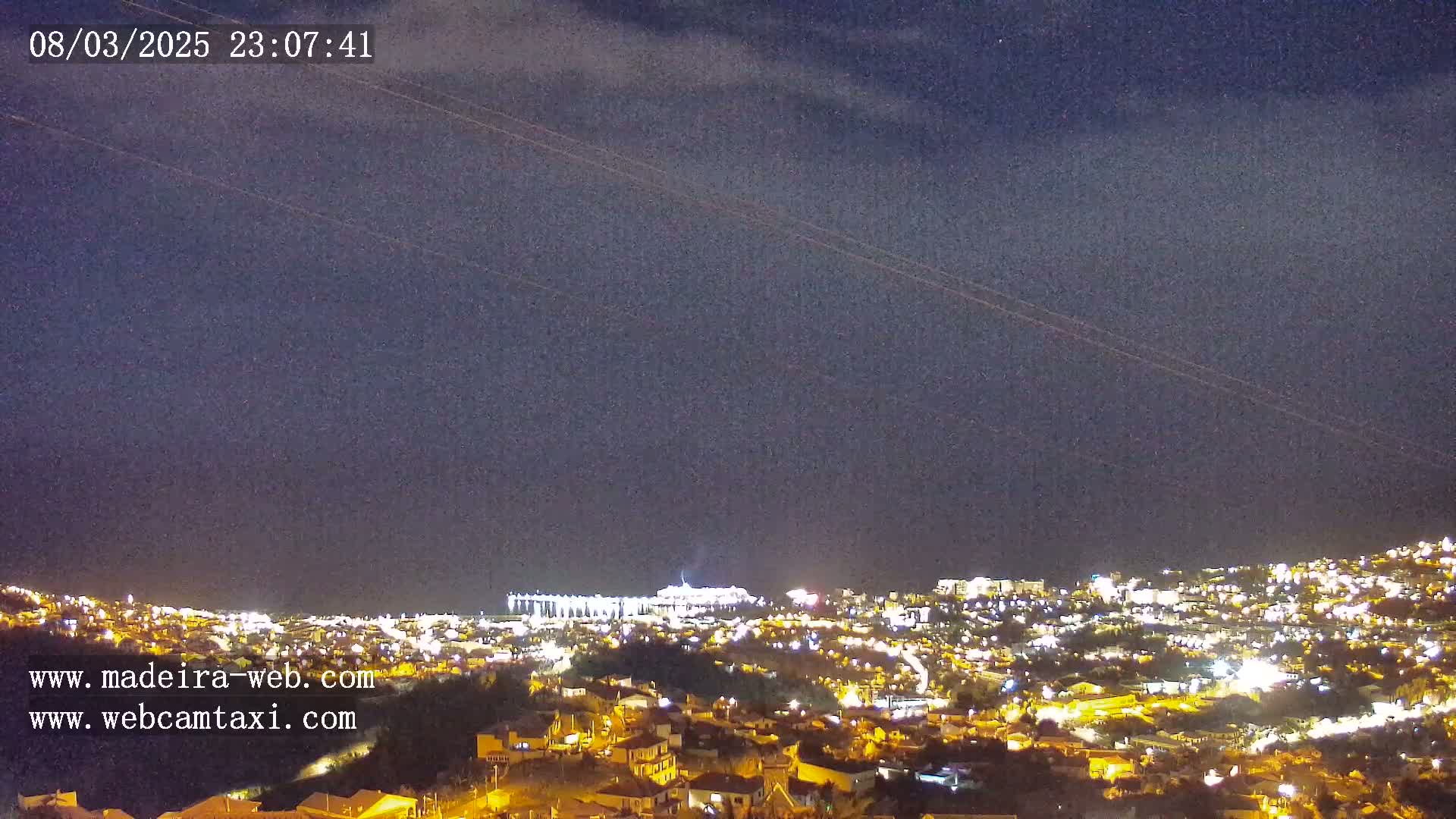 A nighttime view of a city illuminated by lights under a dark, clear sky shows two airplane trails.