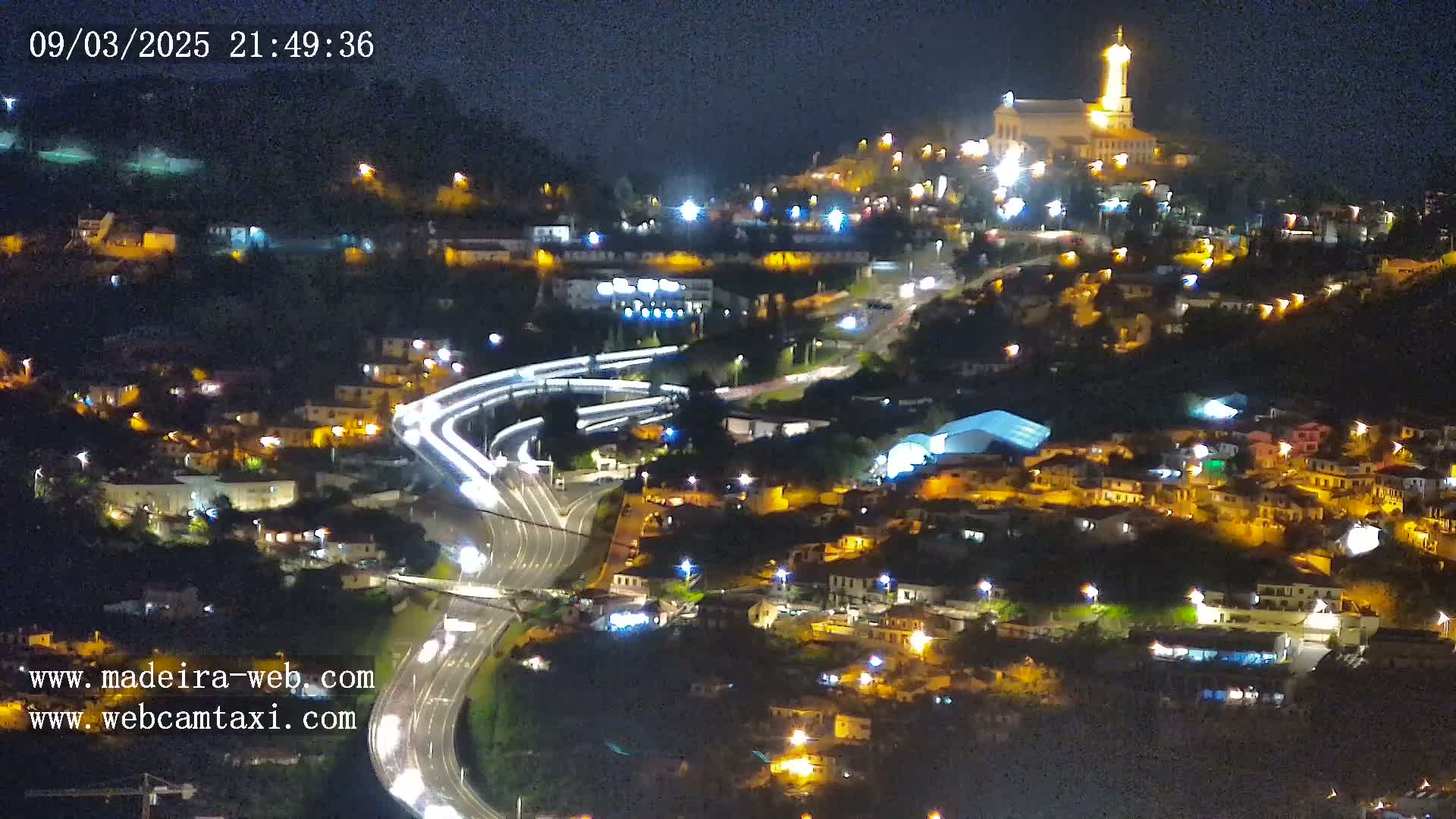 A nighttime aerial view of a town nestled on a hillside, illuminated by streetlights, with a brightly lit church atop the hill and winding roads with light trails below.