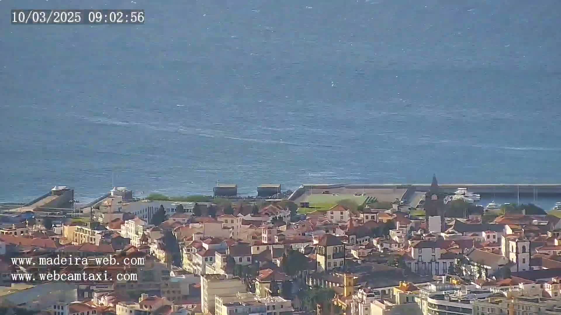 A coastal town with red-roofed buildings sits on a hillside overlooking a calm blue ocean under a clear sky.
