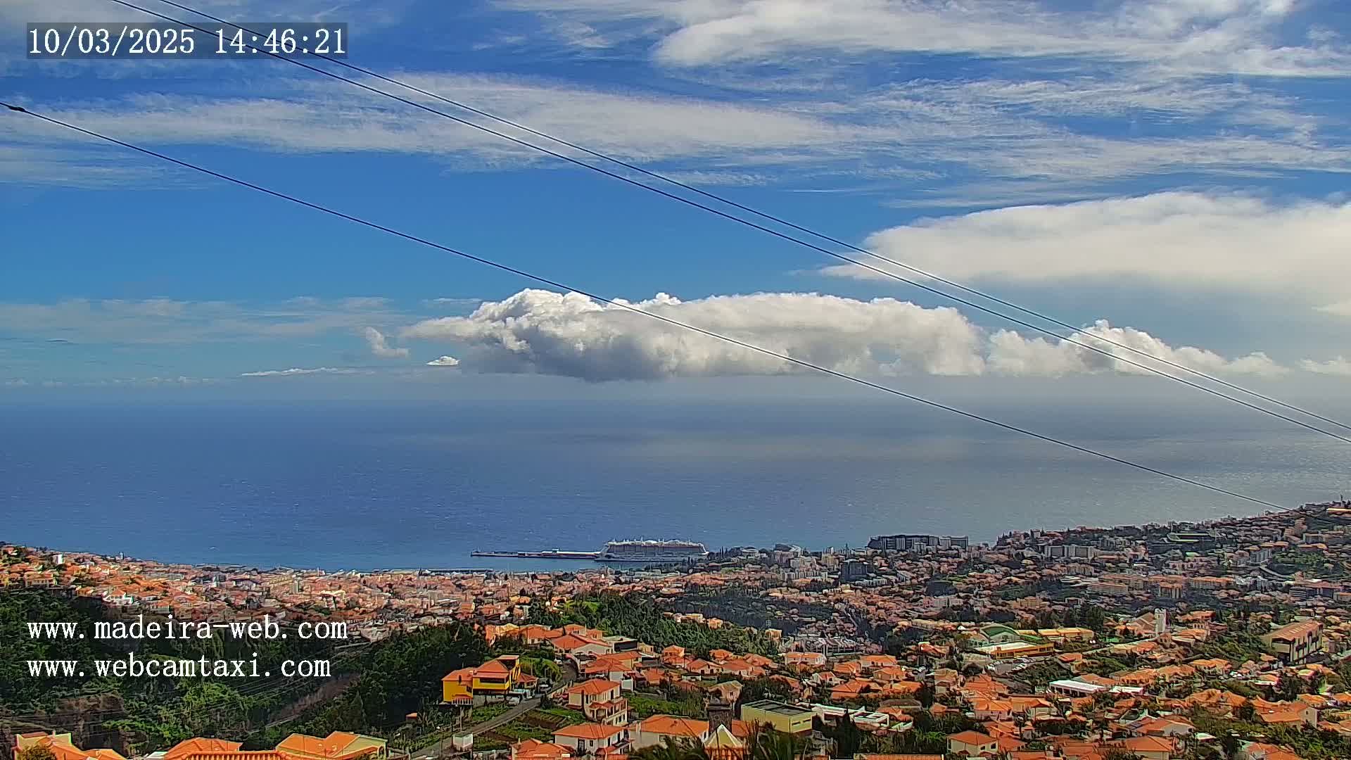 A coastal town with orange-roofed houses stretches toward a calm ocean under a partly cloudy sky with a prominent, puffy cloud formation.