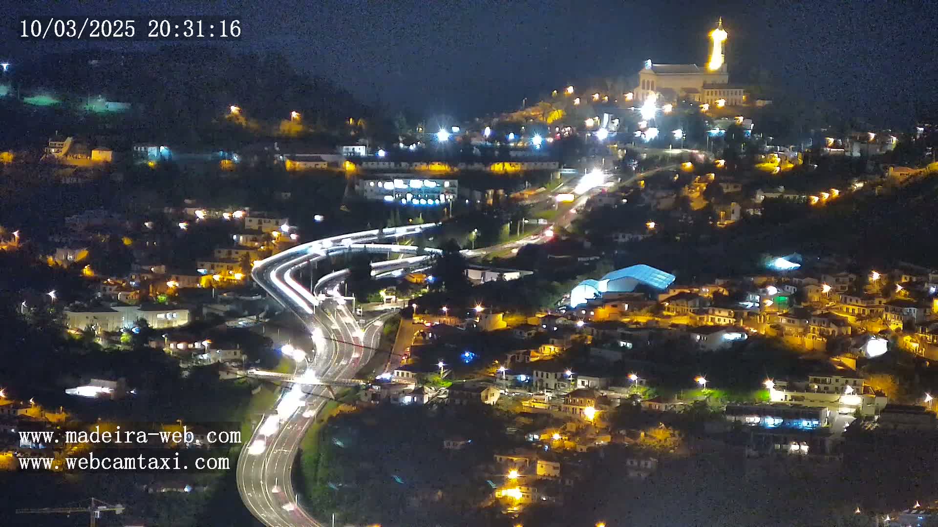 A nighttime aerial view of a city built on a hillside, illuminated by streetlights and featuring a brightly lit building atop a hill, with roads curving through the settlement.