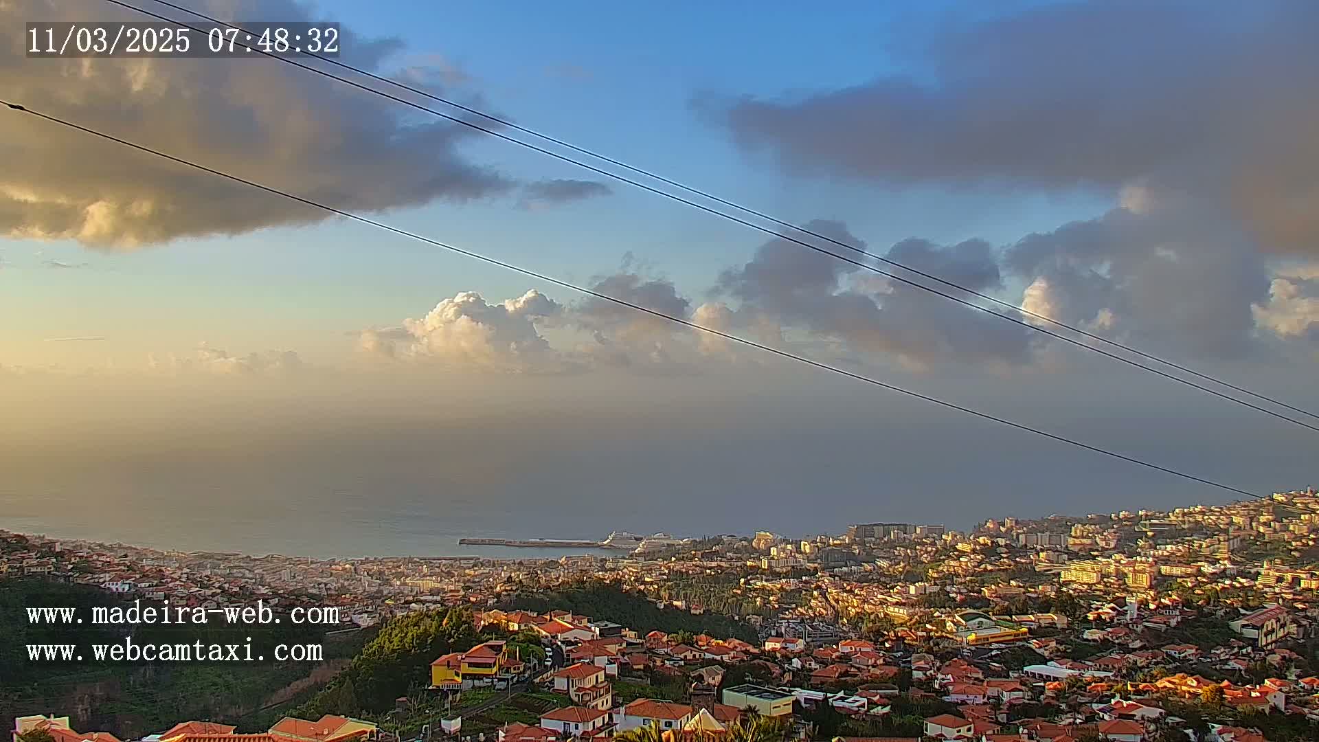 A coastal city with red-roofed houses stretches below a partly cloudy sky at sunrise, with a cruise ship visible in the harbor.