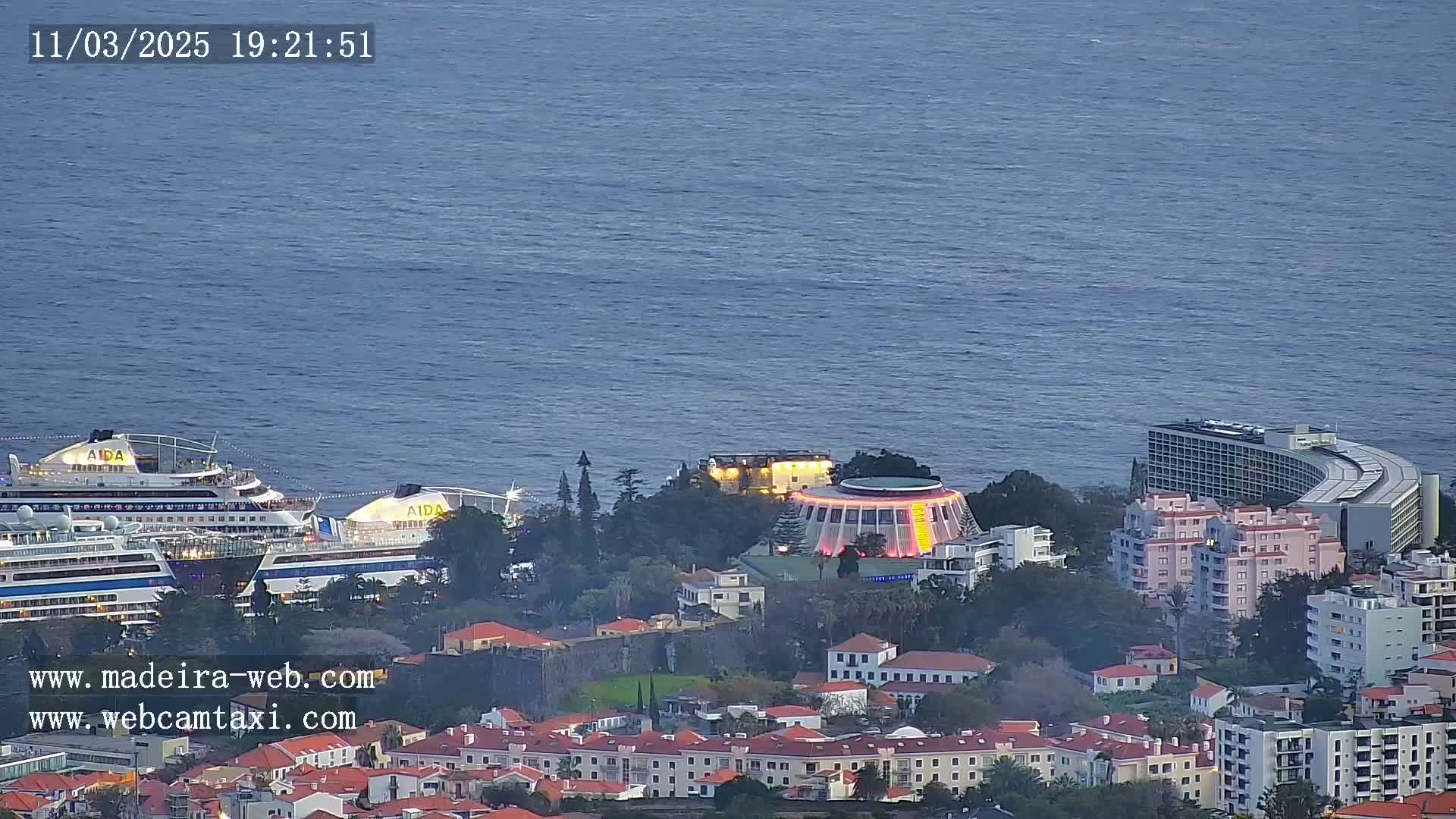 Two cruise ships are docked near a coastal town at dusk, with calm ocean waters in the background.