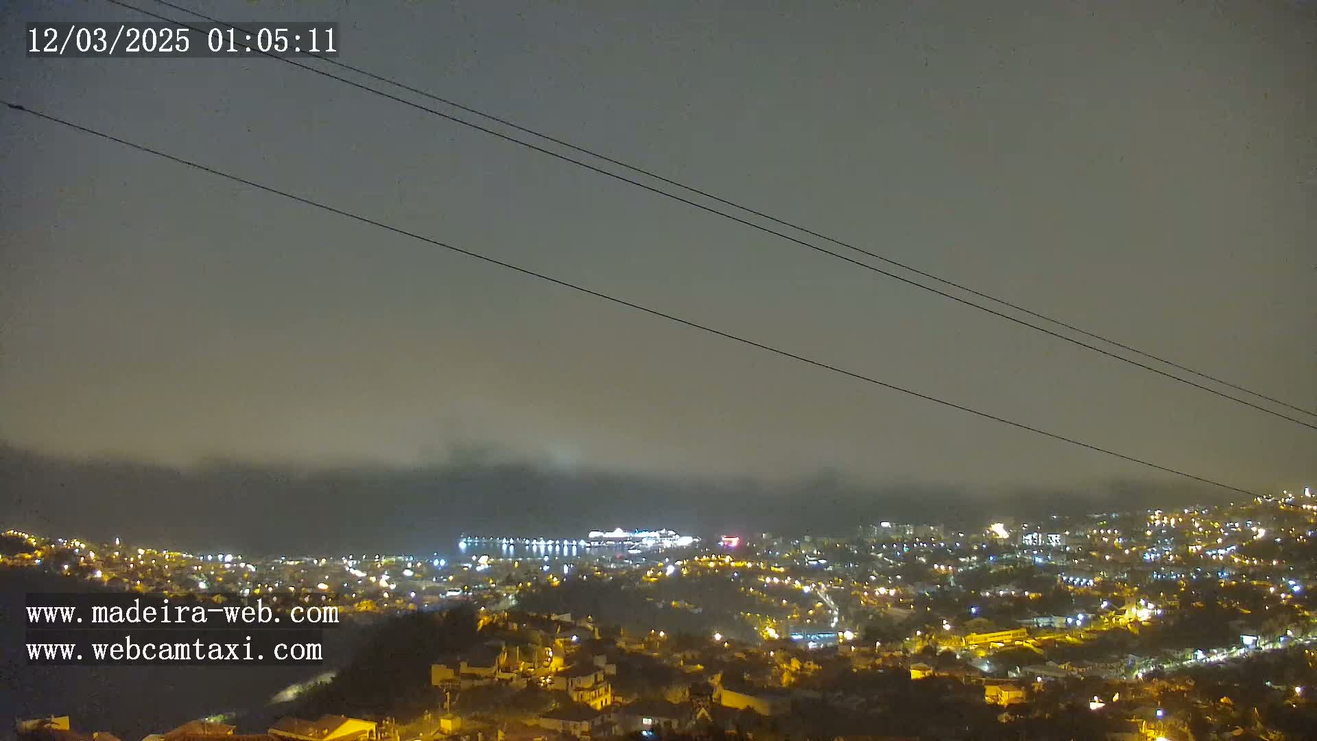A nighttime, foggy view of a coastal city with many illuminated buildings and a large ship visible in the harbor.
