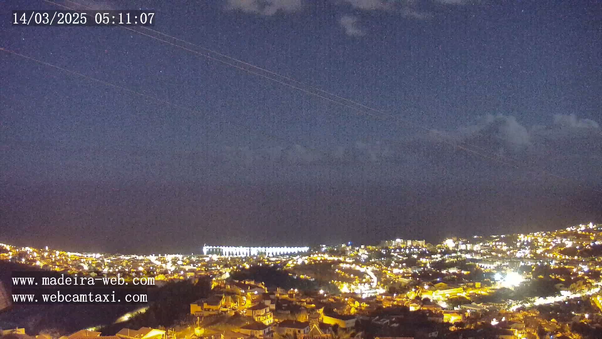 A nighttime view of a city illuminated by lights, under a mostly clear, dark blue sky with a few clouds.