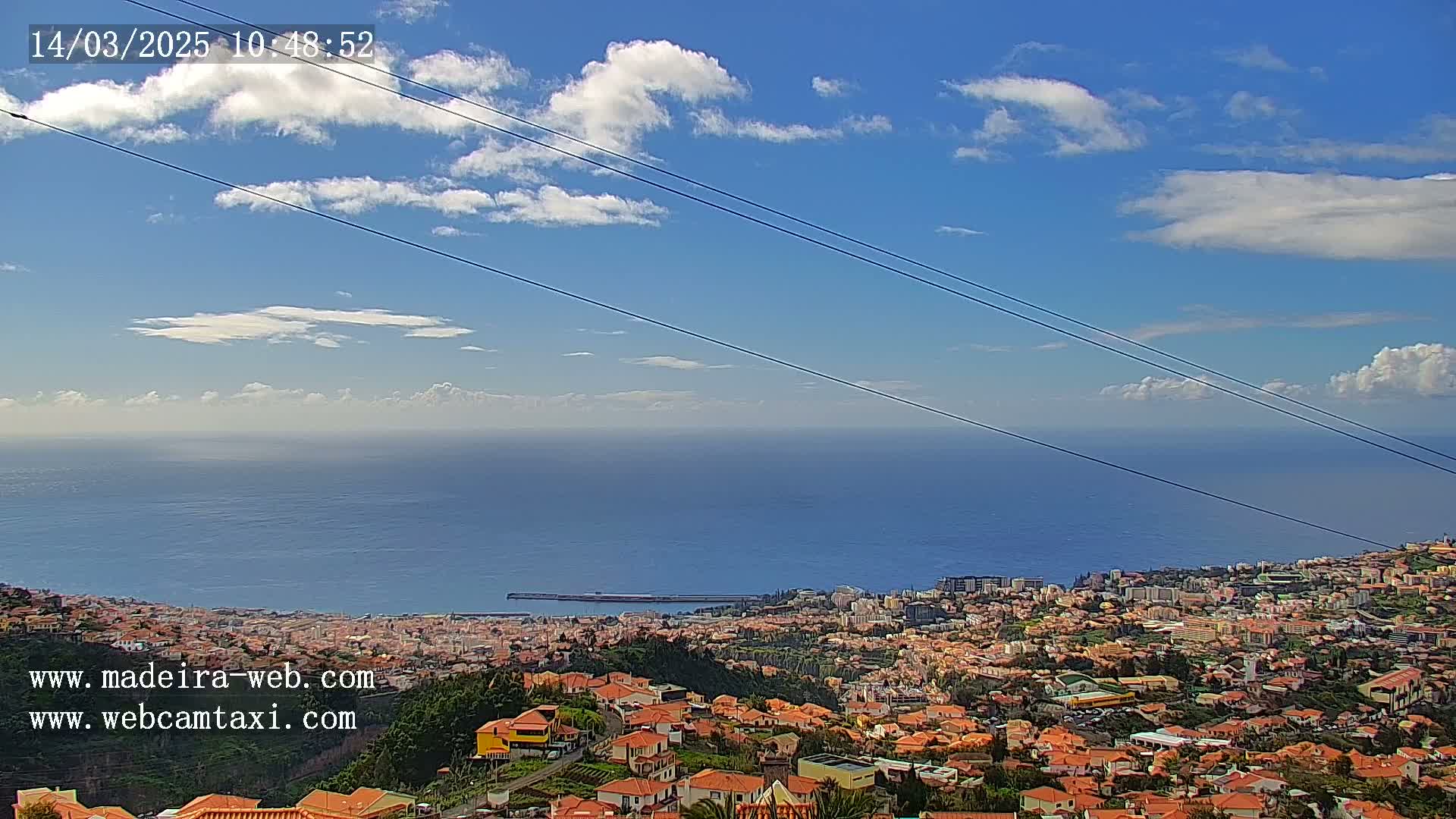 A coastal city with numerous terracotta-roofed buildings stretches toward a calm ocean under a mostly clear, bright blue sky with scattered cumulus clouds.