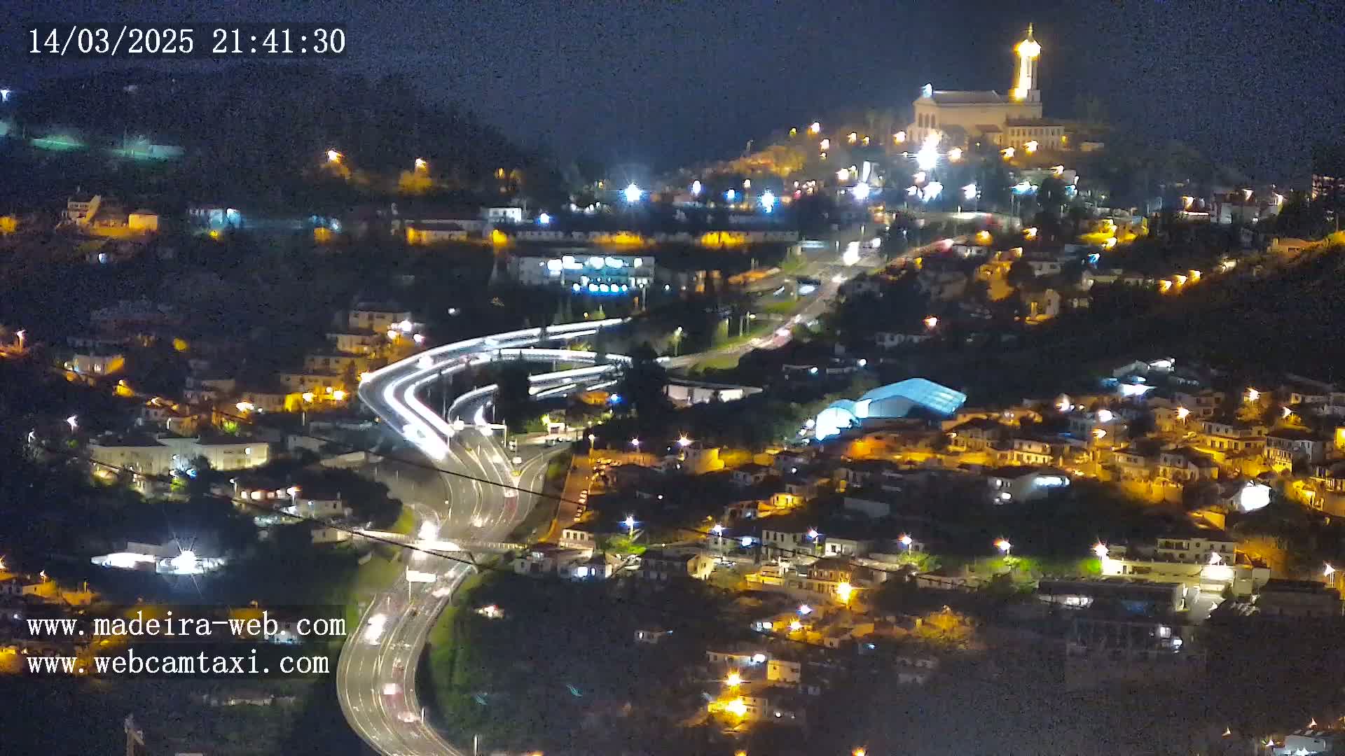 A nighttime aerial view of a town on a hillside, illuminated by streetlights, with a church atop the hill and a highway curving through the landscape.
