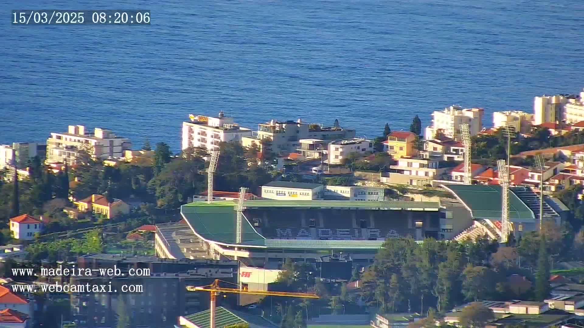 A coastal town with a stadium and residential buildings is visible on a sunny day, with a calm ocean in the background.
