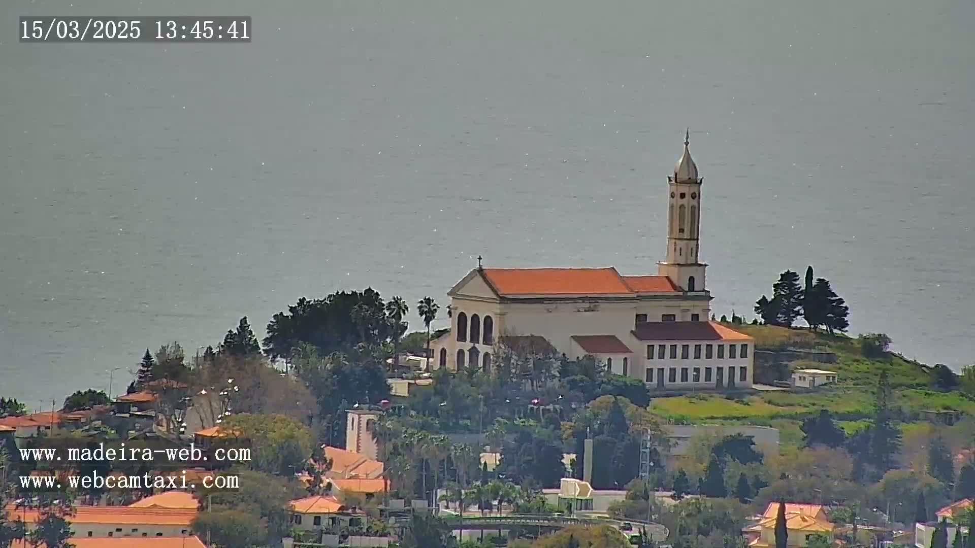 A church with an orange roof and tall bell tower sits atop a hill overlooking a calm body of water under a hazy sky.