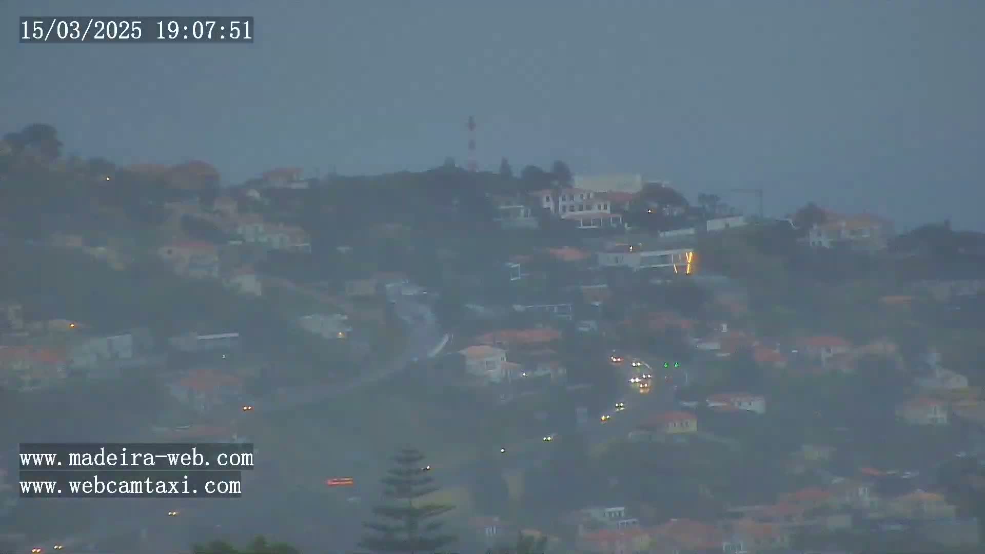 A hazy, twilight view shows a hillside town with scattered lights and a tall structure resembling a tower in the distance.