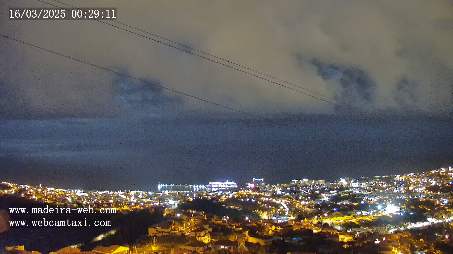 A nighttime view of a coastal city with lights illuminating buildings, a cruise ship visible in the harbor, and a cloudy sky above.
