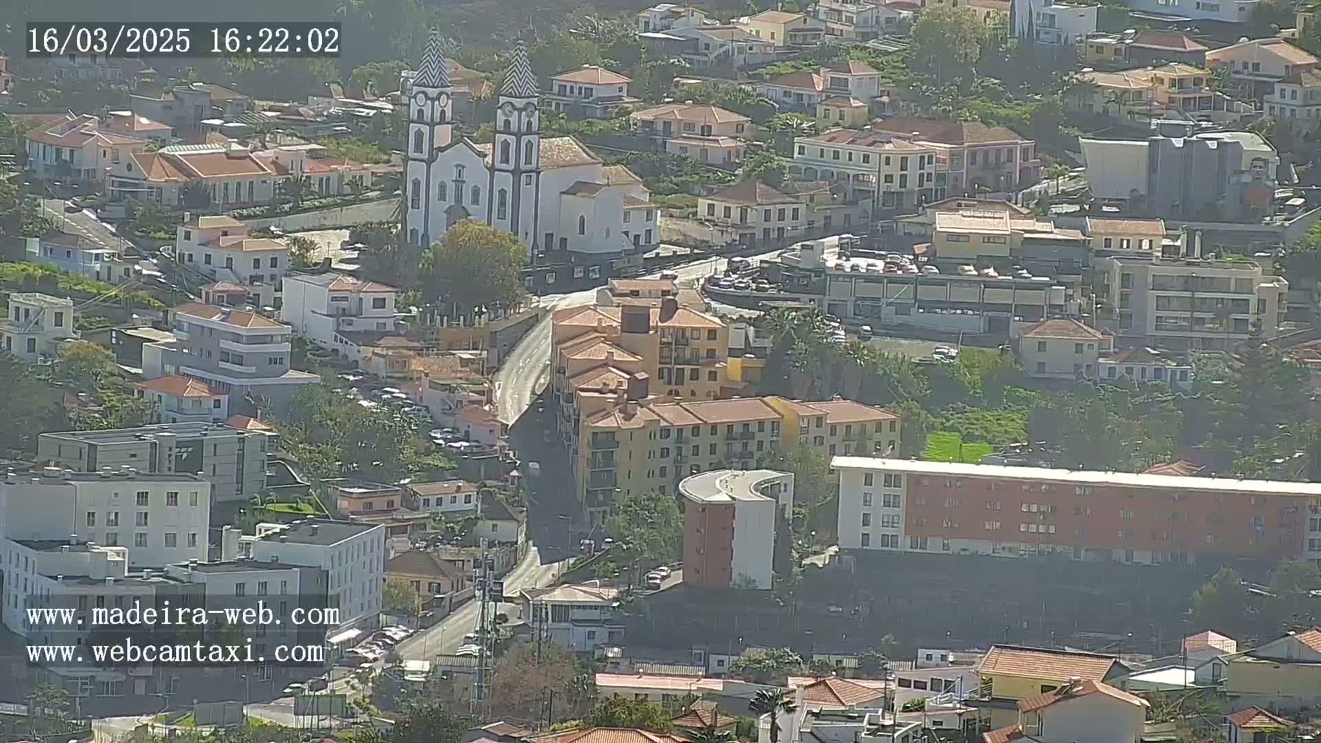 An aerial view of a town on a sunny day, featuring a white church with twin towers and numerous buildings, some modern and some appearing older.