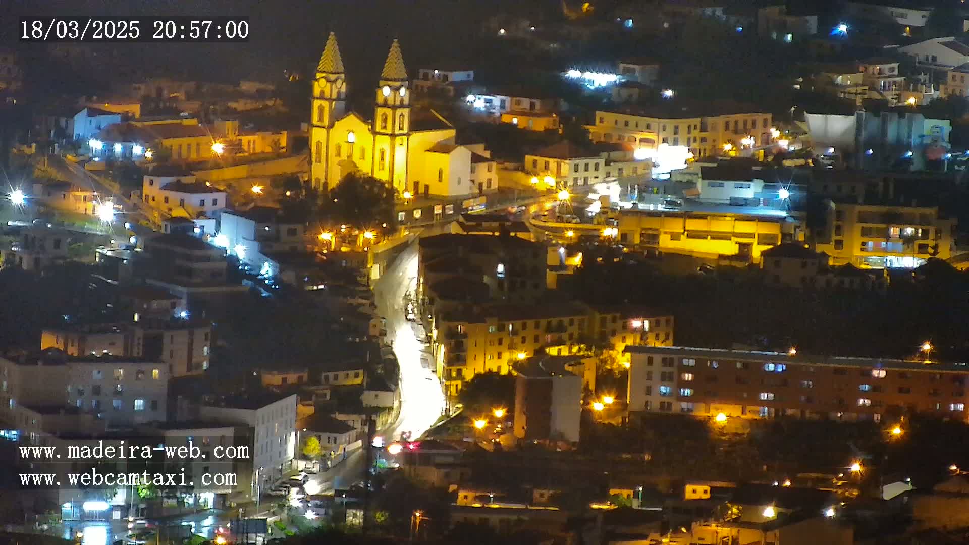 A nighttime view of a town, illuminated by streetlights, featuring a prominent church with twin towers, and winding streets.