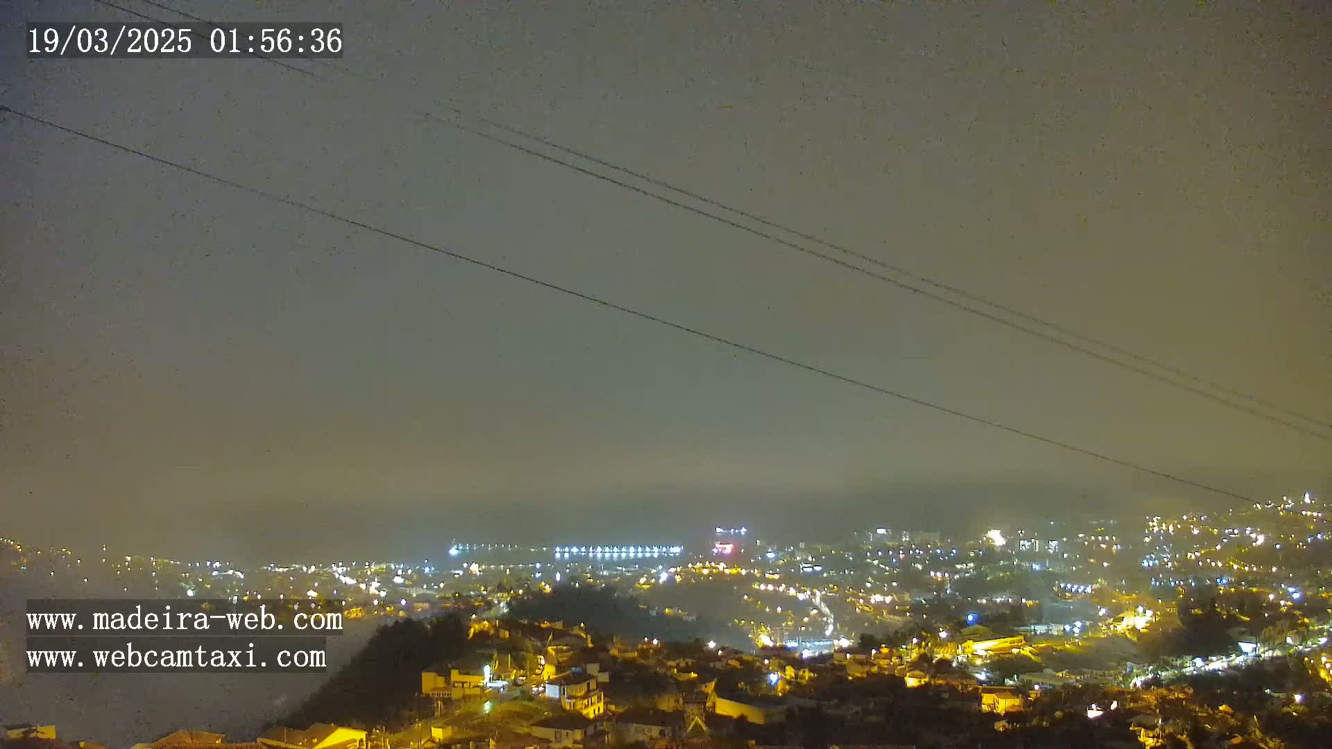 A nighttime, foggy view overlooking a city illuminated with streetlights, extending to a coastline under a dark sky.